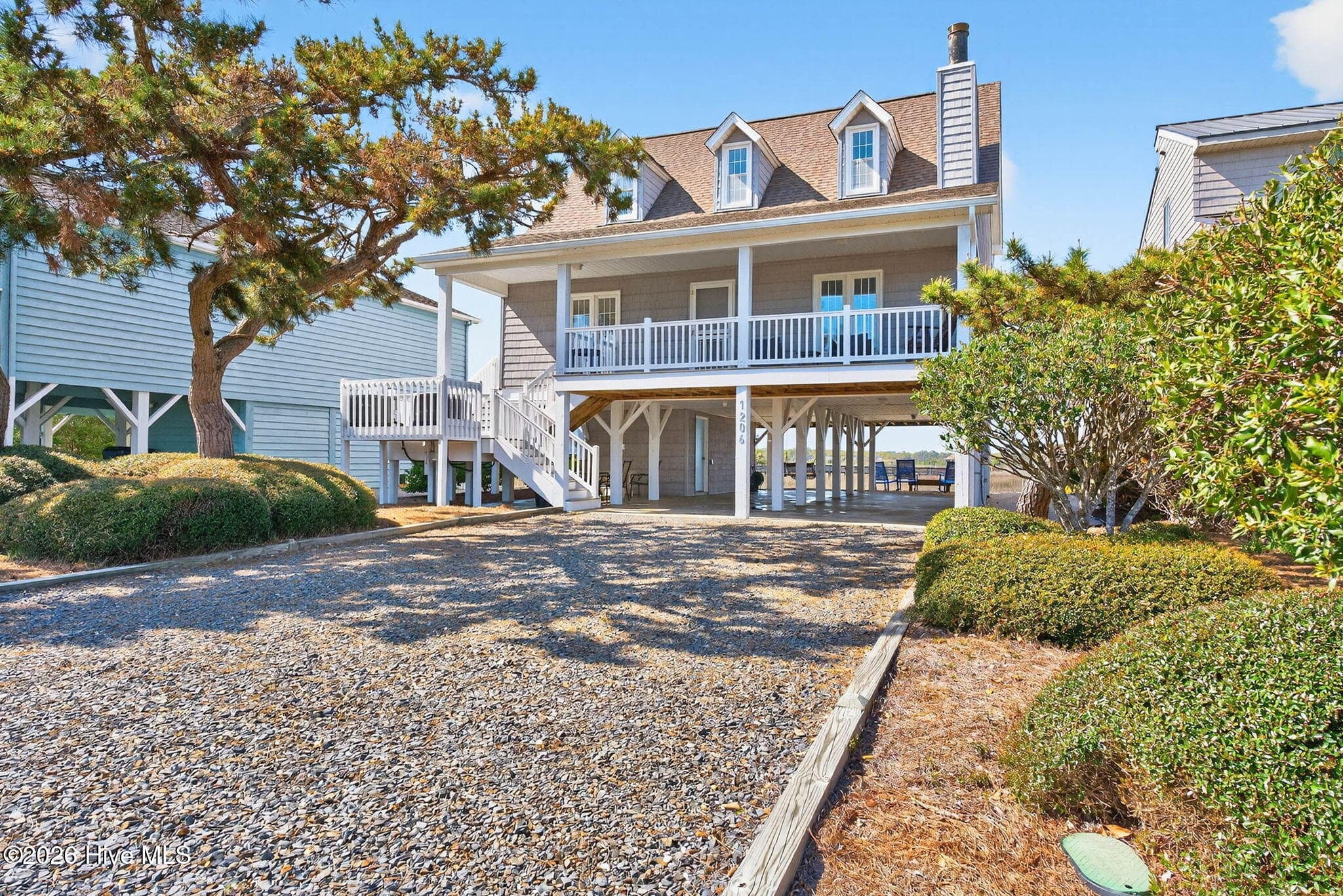 Spacious gravel driveway leading to elevated beach home with covered parking and classic coastal architecture. - 1206 N Shore Drive W, Sunset Beach, NC 28468
