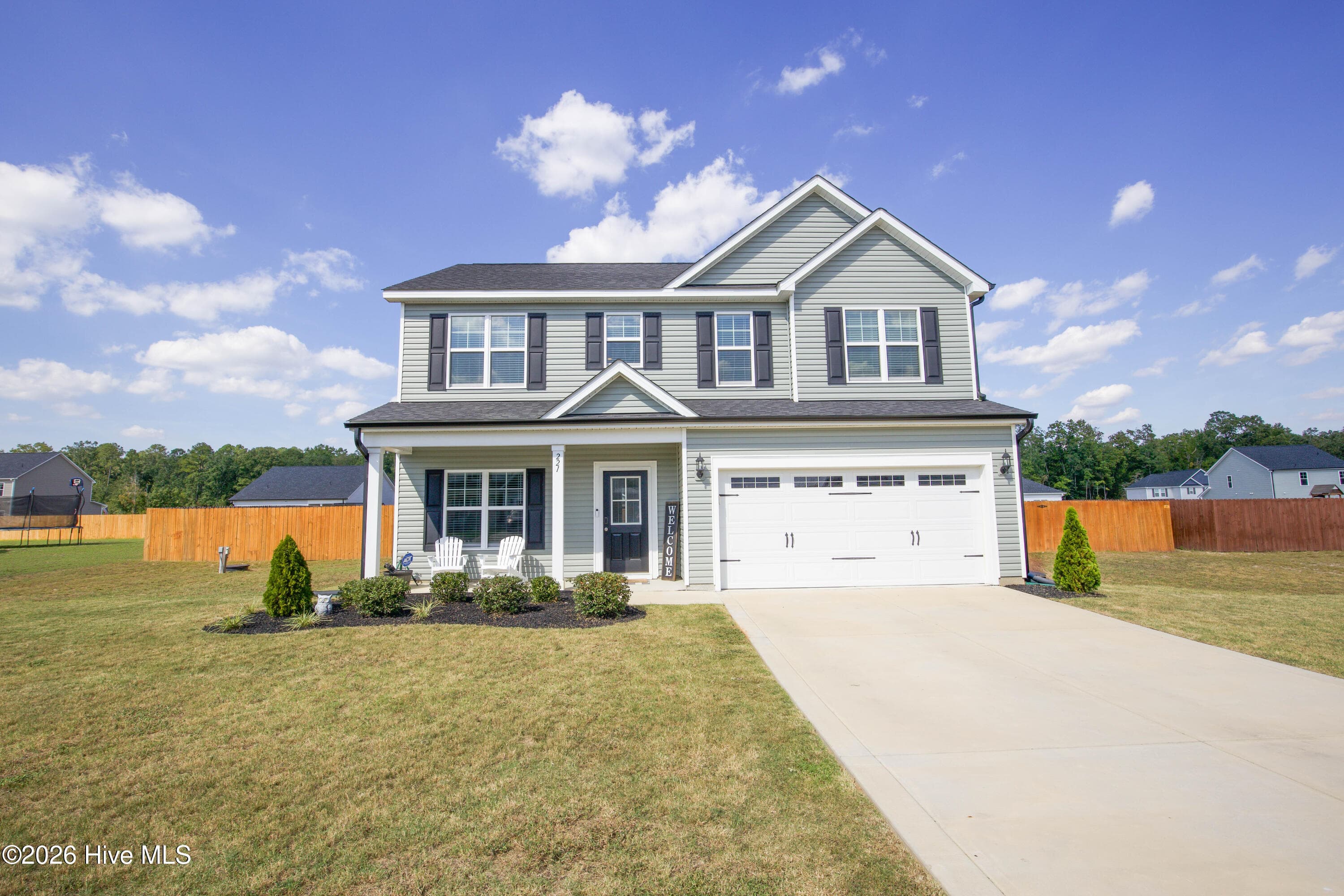 This straight-on view of the front of 221 Vega Lane shows how the home is set back from the street with an open front lawn and a long concrete driveway. Shutters, a gabled roofline, and vinyl siding give the two-story facade a clean, traditional look. A wood privacy fence is visible on each side of the house, marking where the fenced backyard begins. The house sits well back from the road, and it offers a practical option for buyers searching the Fort Bragg area who want a newer home with yard space. - 221 Vega Lane, Linden, NC 28356