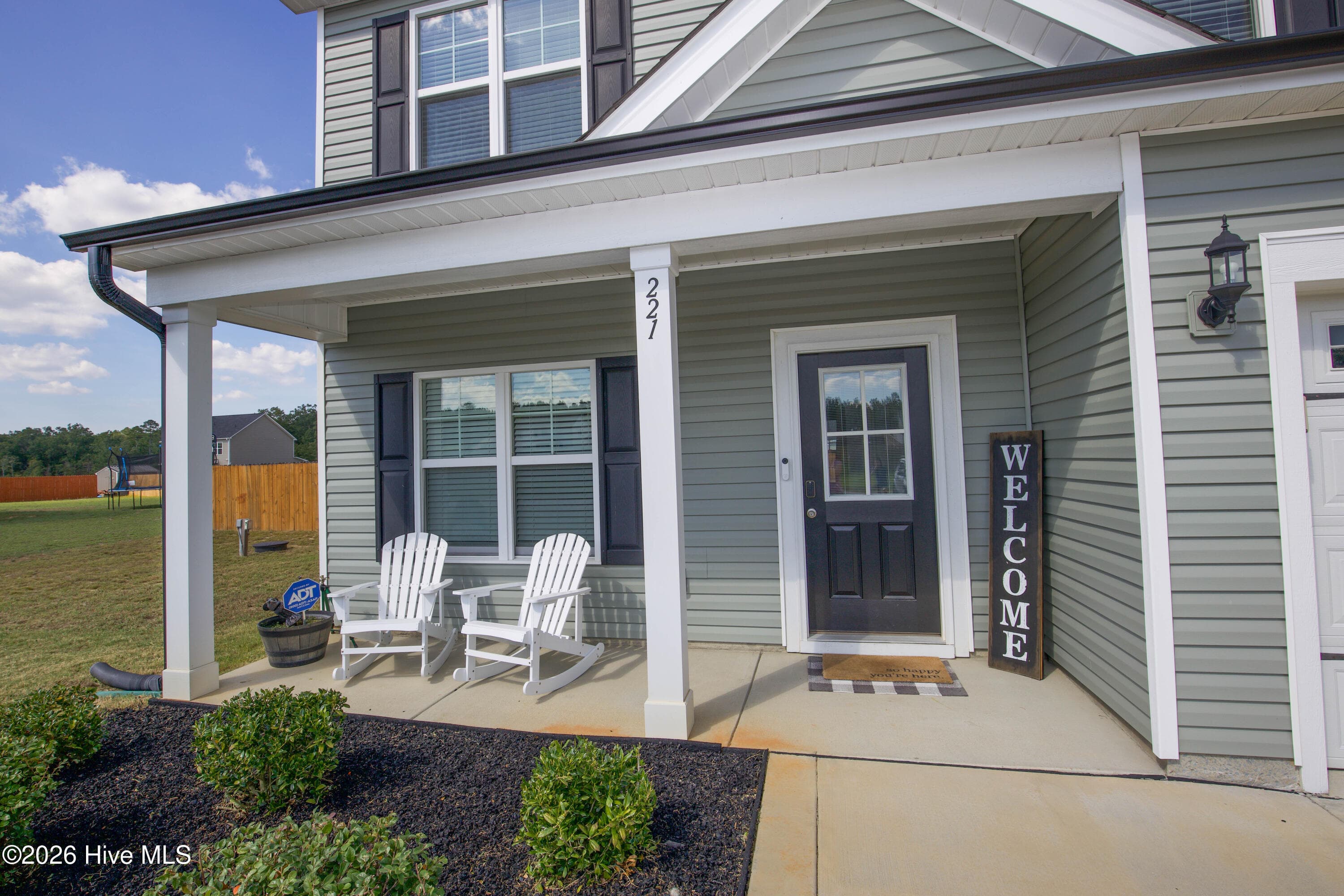 Up close, the front entry shows a half-glass front door, a carriage-style porch light, and the house number on the porch column. The covered porch keeps the doorway sheltered when you are coming and going in the rain. From this door the walkthrough begins, leading into the foyer and the main level of this 2024-built home. - 221 Vega Lane, Linden, NC 28356