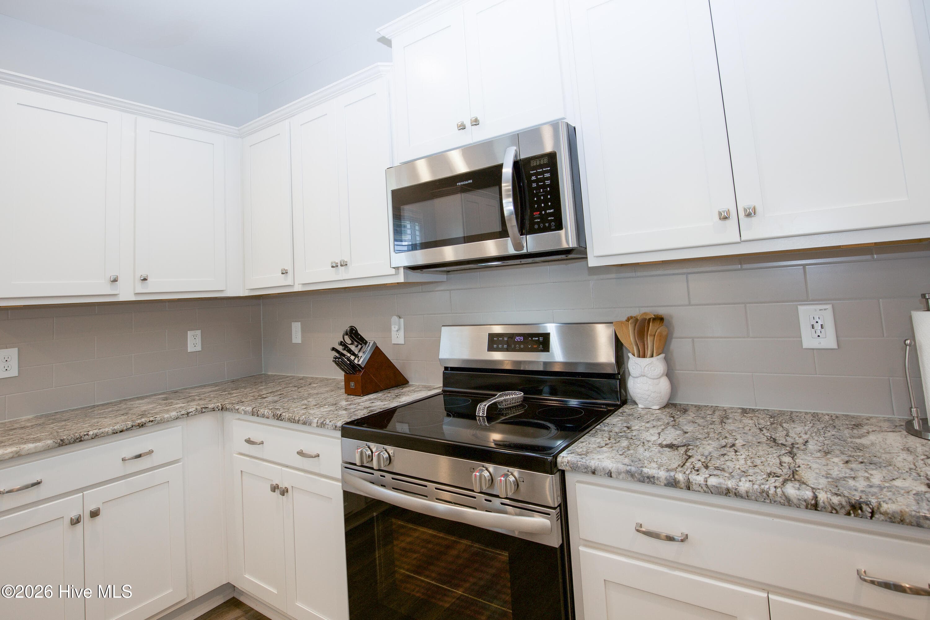 A closer look at the range wall shows the detail in the white shaker-style cabinet doors, brushed-finish hardware, and the tile backsplash running behind the granite counters. Upper cabinets extend along the wall for everyday storage of dishes and pantry items. The corner layout keeps counter space on both sides of the range, so there is room to set down pots and ingredients while you cook. - 221 Vega Lane, Linden, NC 28356