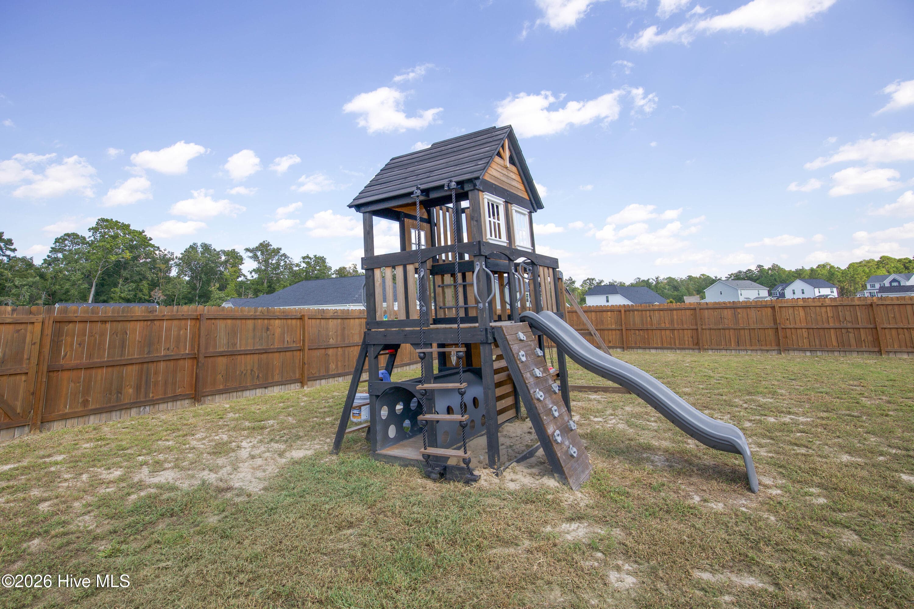 From another angle, the play structure shows its climbing wall, slide, and covered upper deck. Behind it, the wood privacy fence and trees beyond the property line frame the back of the yard. The playground equipment is included in the sale. - 221 Vega Lane, Linden, NC 28356