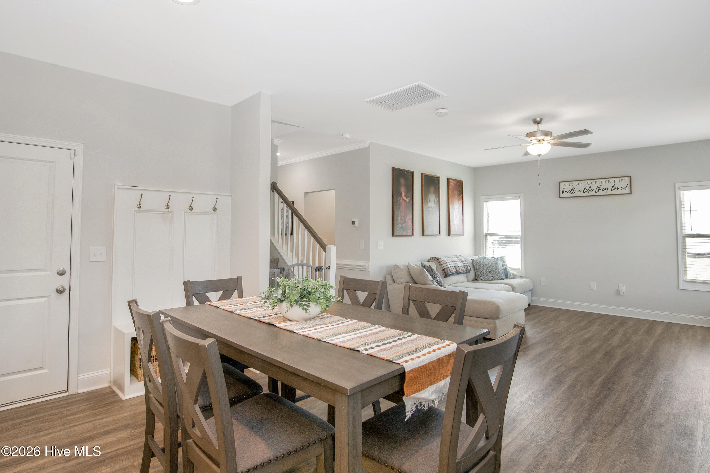 From the end of the dining table, the main level opens toward the Great Room, with the staircase rising in the background. The drop-zone bench and door sit along the left wall, keeping that everyday entry out of the main living space. Wood-look flooring runs continuously from the dining area into the living room, which helps the main level feel like one connected space. - 221 Vega Lane, Linden, NC 28356