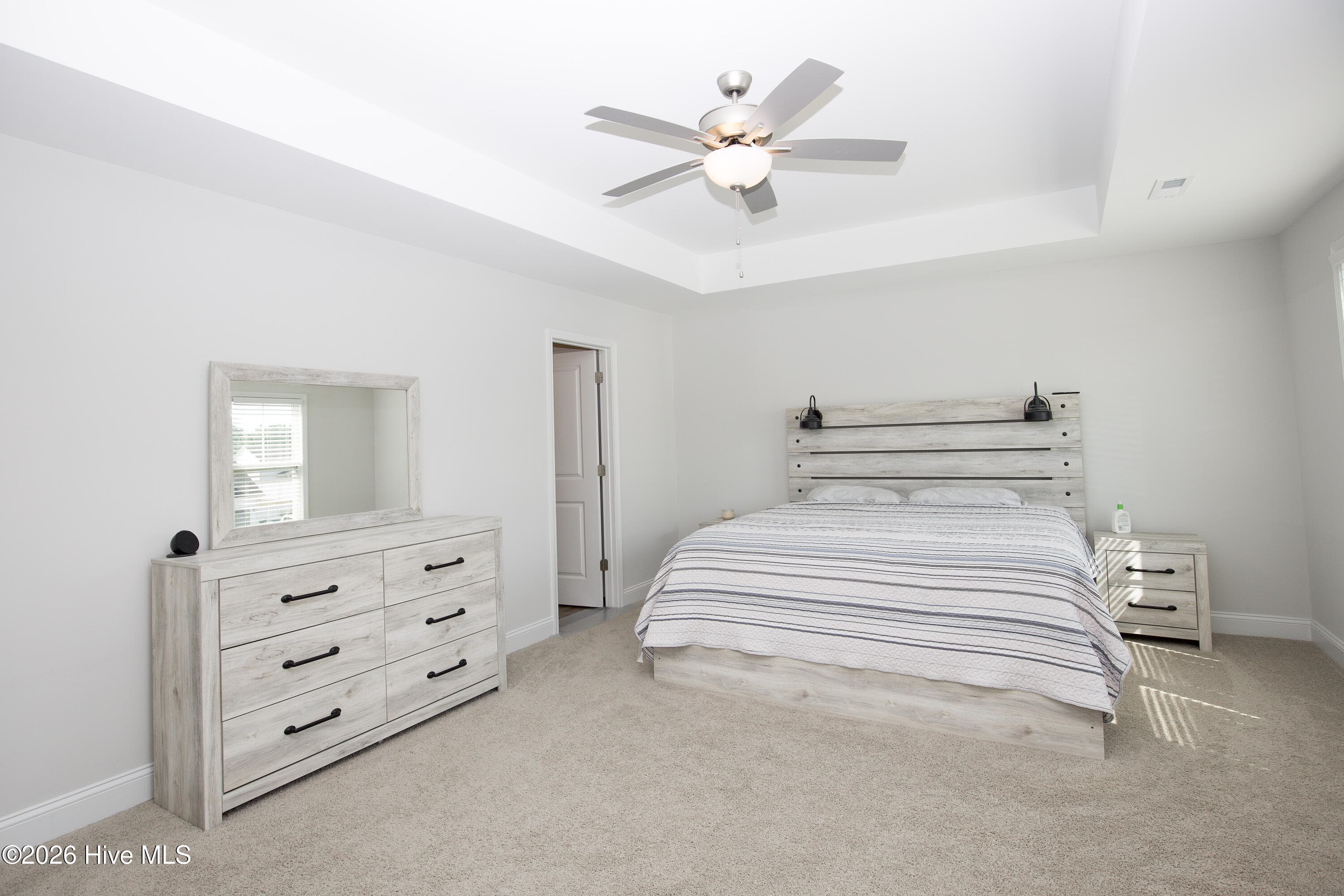 From the far side of the room, this view shows the primary bedroom's layout with the bed on one wall and the doorway to the bath and closet on the other. The tray ceiling and ceiling fan are centered over the sleeping area. Carpet and neutral walls keep the room ready for your own furniture and finishes. - 221 Vega Lane, Linden, NC 28356