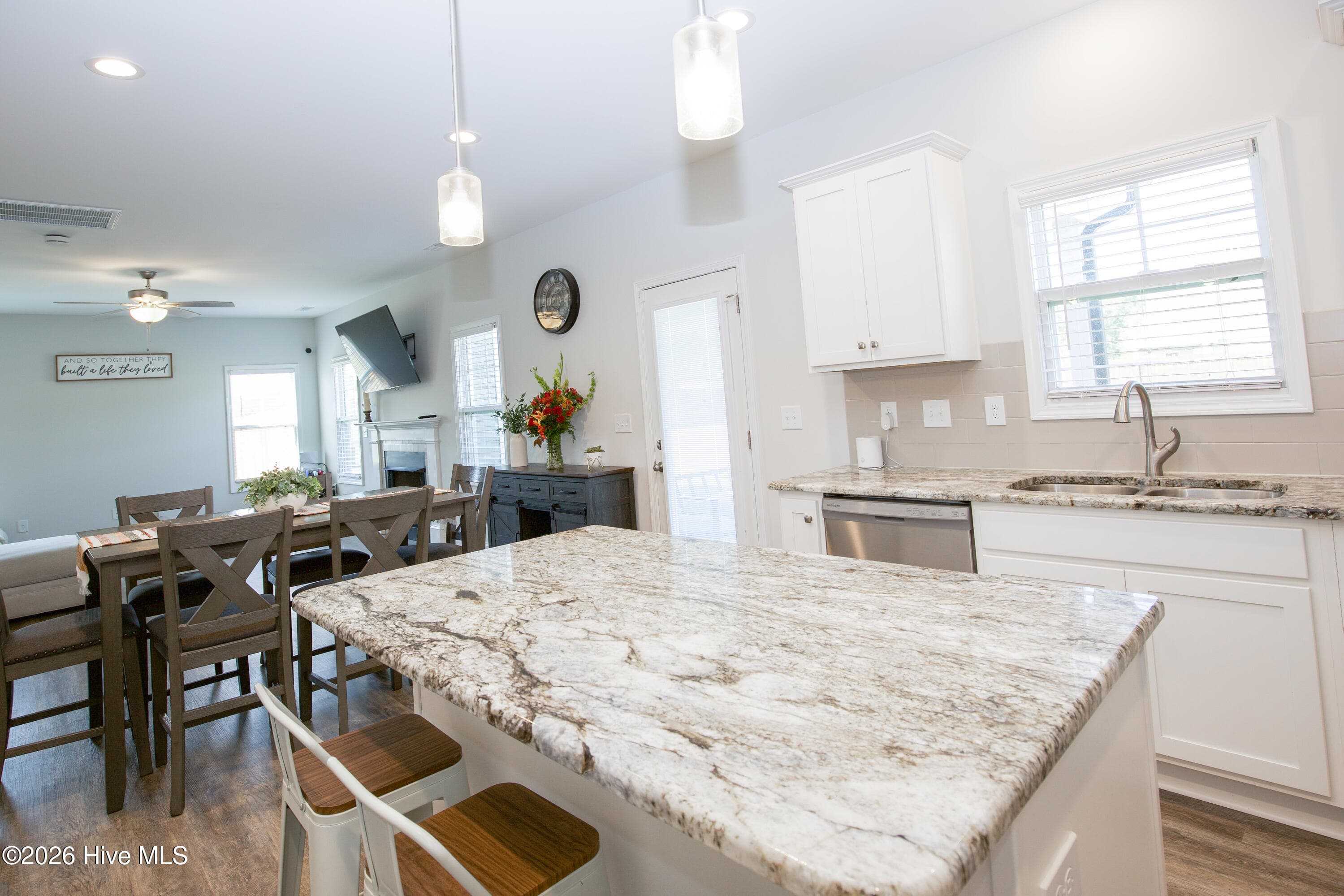 From the sink side of the kitchen, the granite island leads your eye toward the dining table and the Great Room fireplace beyond. Windows along the back wall bring in daylight across the whole main level. The rear door to the covered patio is visible here as well, linking the kitchen to the backyard. - 221 Vega Lane, Linden, NC 28356