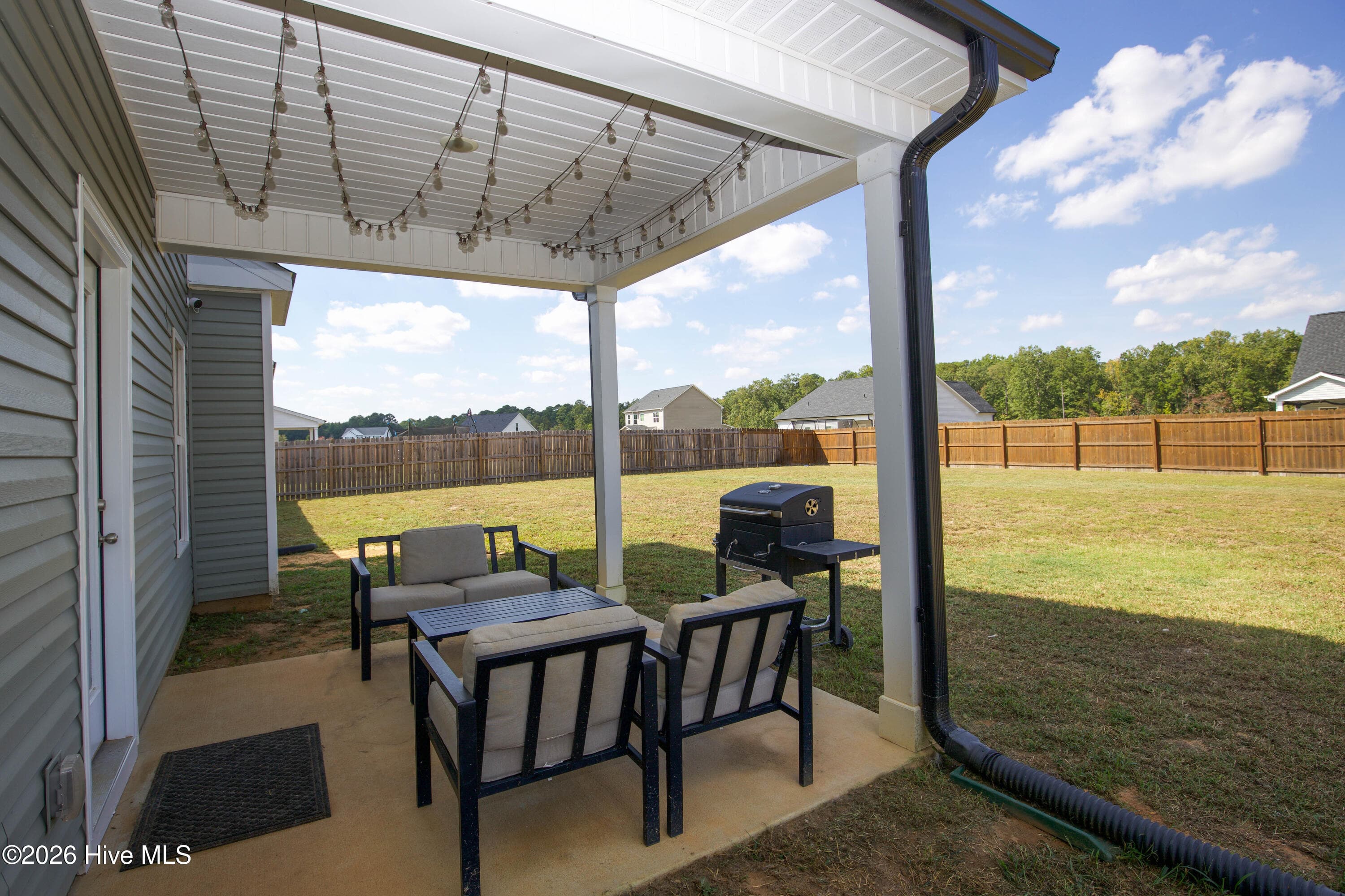 Off the kitchen, the covered rear patio provides a shaded spot for outdoor seating and a grill, with string lights hung under the roof. The patio looks out over the fenced, mostly level backyard. According to the floor plan, it measures approximately 10 feet by 10 feet with a 9'6" ceiling. - 221 Vega Lane, Linden, NC 28356