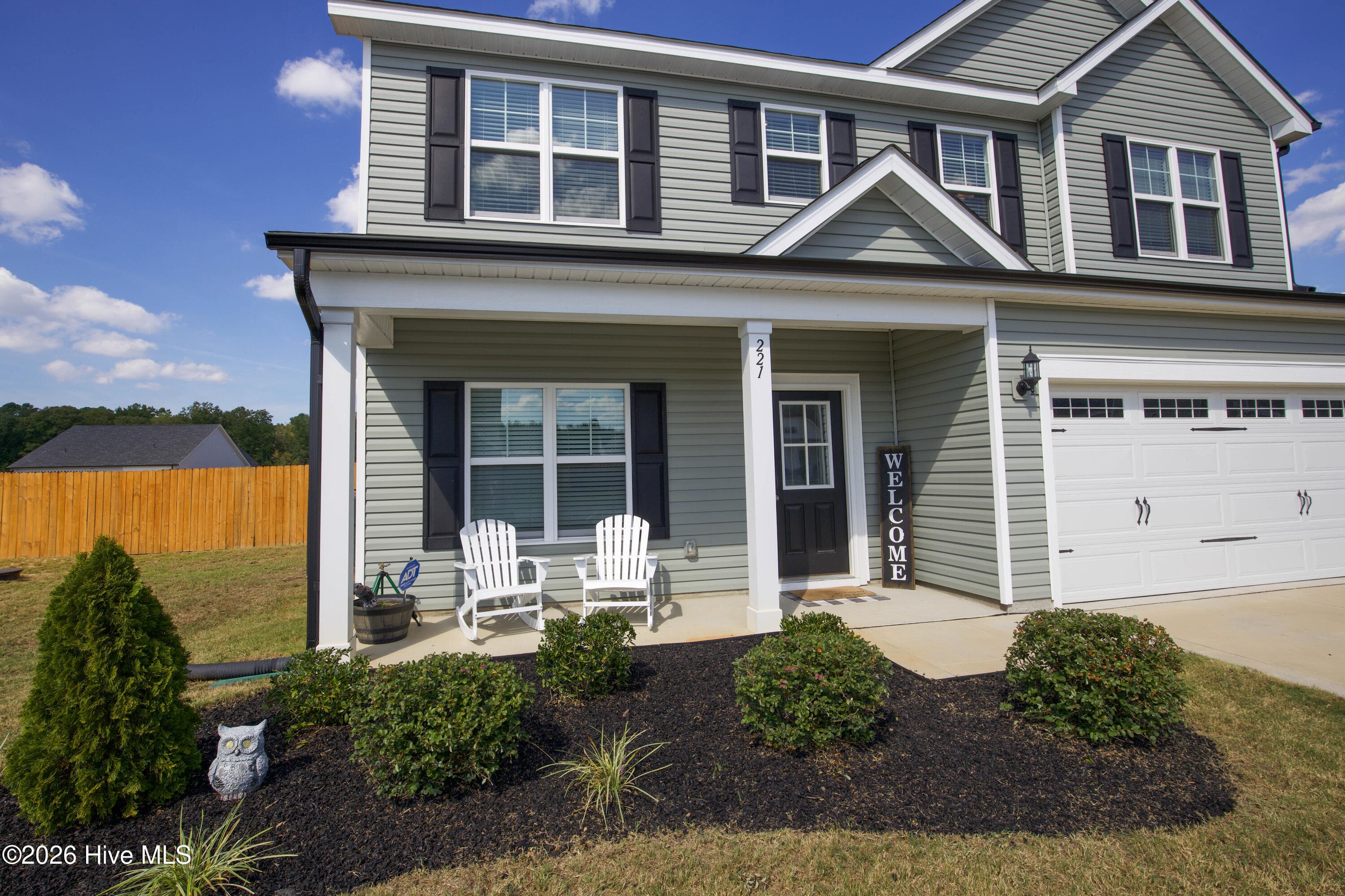 A covered front porch runs across the front of the house beside the garage, with room for a pair of chairs and a view out over the yard. Mulched planting beds with shrubs frame the entry and soften the front of the home. White porch columns and a dark front door with a glass panel give the entry a finished look. The porch is a simple, practical place to sit outside in the morning or welcome guests. - 221 Vega Lane, Linden, NC 28356