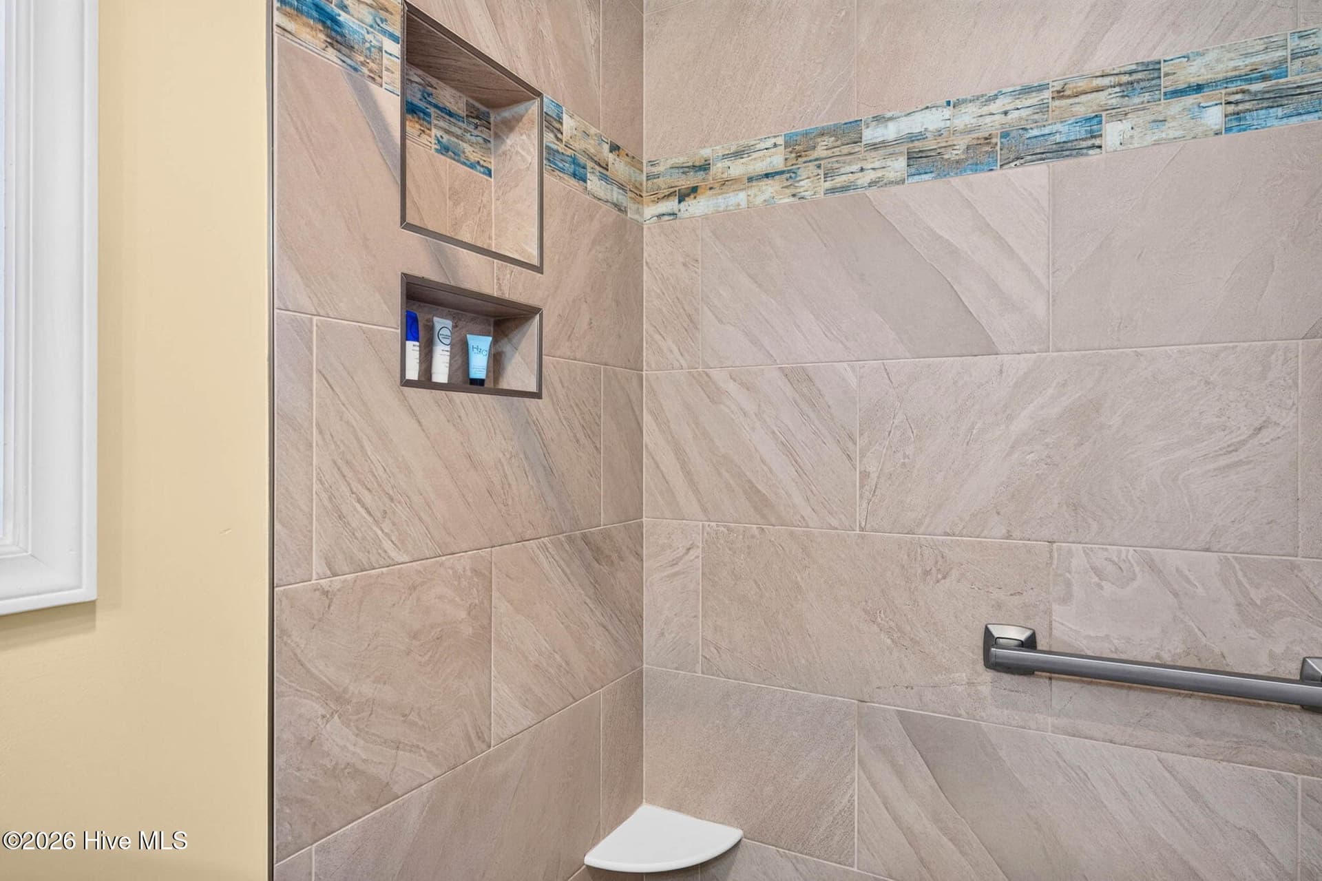 Detailed view of the primary suite's custom tiled walk-in shower with built-in storage and grab bars. - 1650 Frink Street SW, Ocean Isle Beach, NC 28469