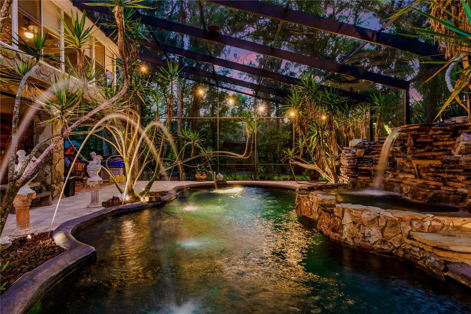 A tropical screened pool retreat surrounded by mature landscaping features dramatic rock waterfalls, ambient lighting, and a resort-inspired setting designed for relaxing and entertaining at home. - 11526 RIVER COUNTRY DR, RIVERVIEW