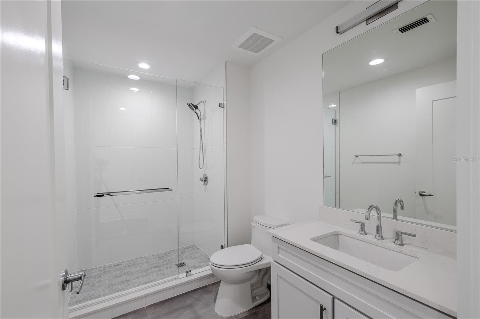 Bathroom 2 offers a sleek, contemporary design with a frameless glass-enclosed walk-in shower featuring large-format white tile and a mosaic tile shower floor. A quartz-topped vanity with under-mount sink, polished chrome fixtures, and a wide mirror with integrated lighting create a bright, spa-like atmosphere. Clean lines and neutral tones make this en-suite both elegant and functional—perfect for guests or as a private bath for the secondary bedroom. - 920 N OSCEOLA AVE #703, CLEARWATER