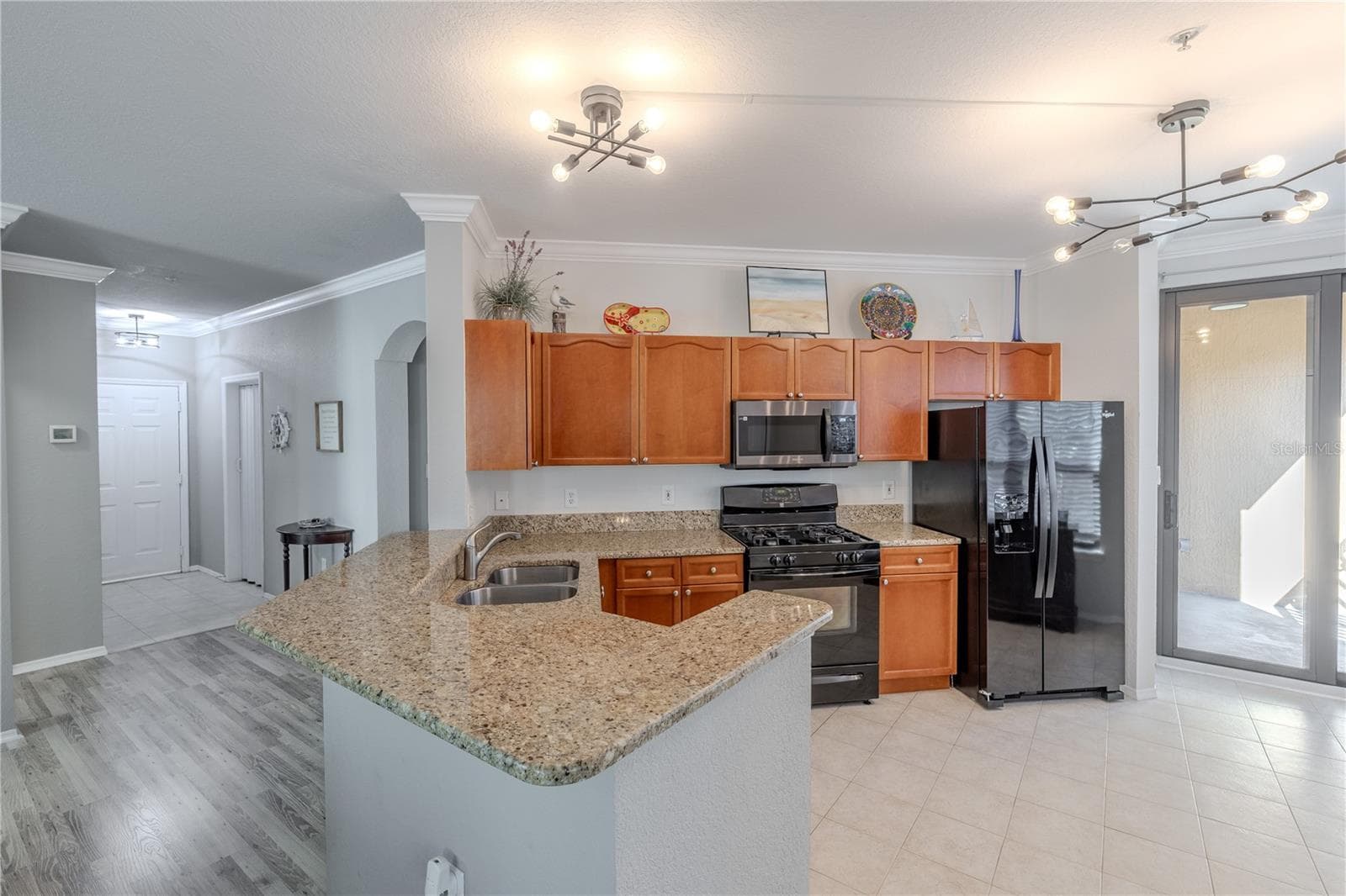 A well-designed kitchen combines expansive granite counters, a generous breakfast bar, warm wood cabinetry, stainless appliances, and modern lighting, all opening seamlessly to the private balcony for effortless indoor–outdoor living. - 2738 VIA TIVOLI #220B, CLEARWATER
