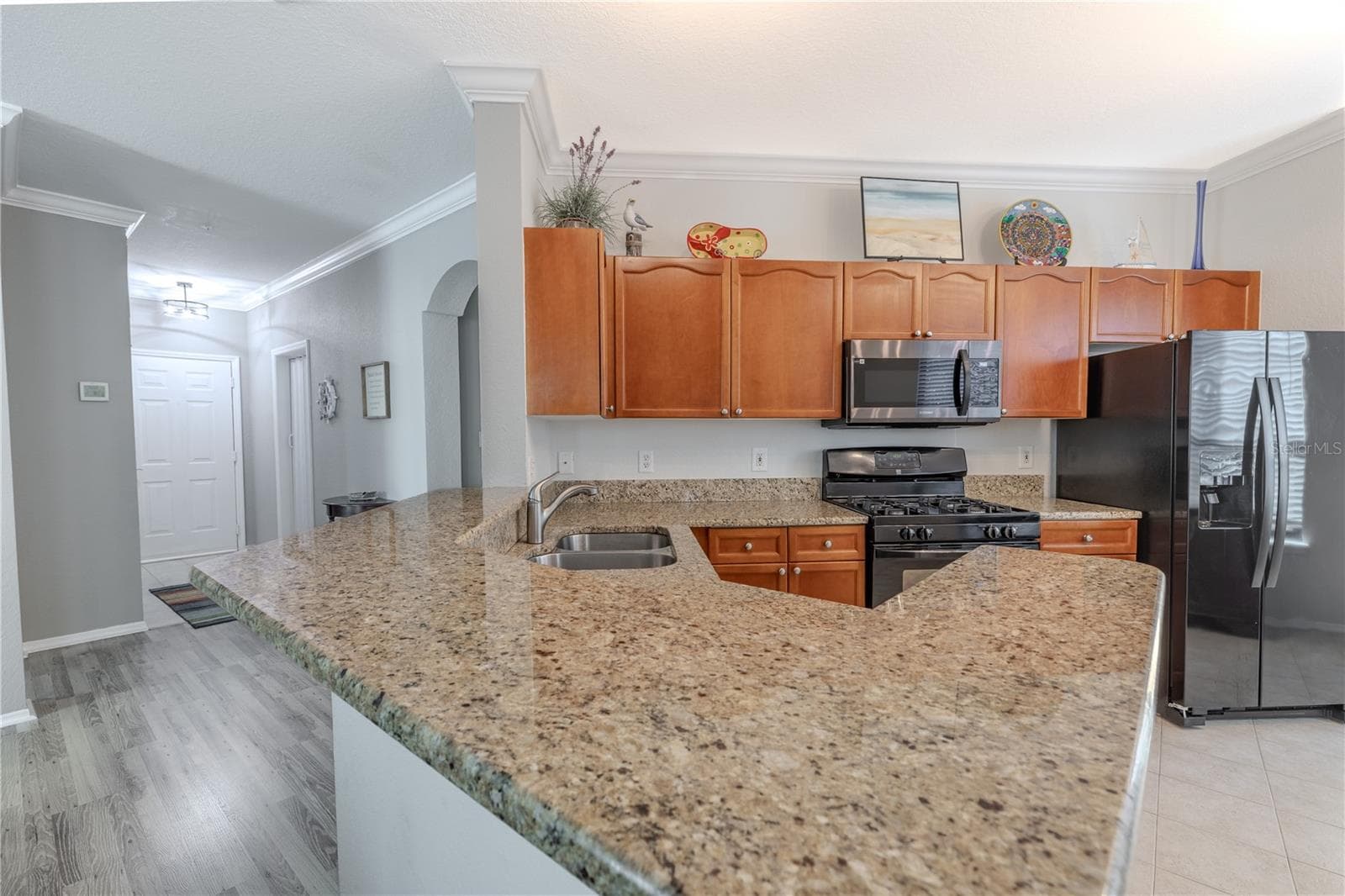 This angle highlights the kitchen’s efficient layout with granite countertops, warm wood cabinetry, stainless appliances, and a spacious breakfast bar that opens directly to the living area, enhancing both functionality and flow. - 2738 VIA TIVOLI #220B, CLEARWATER