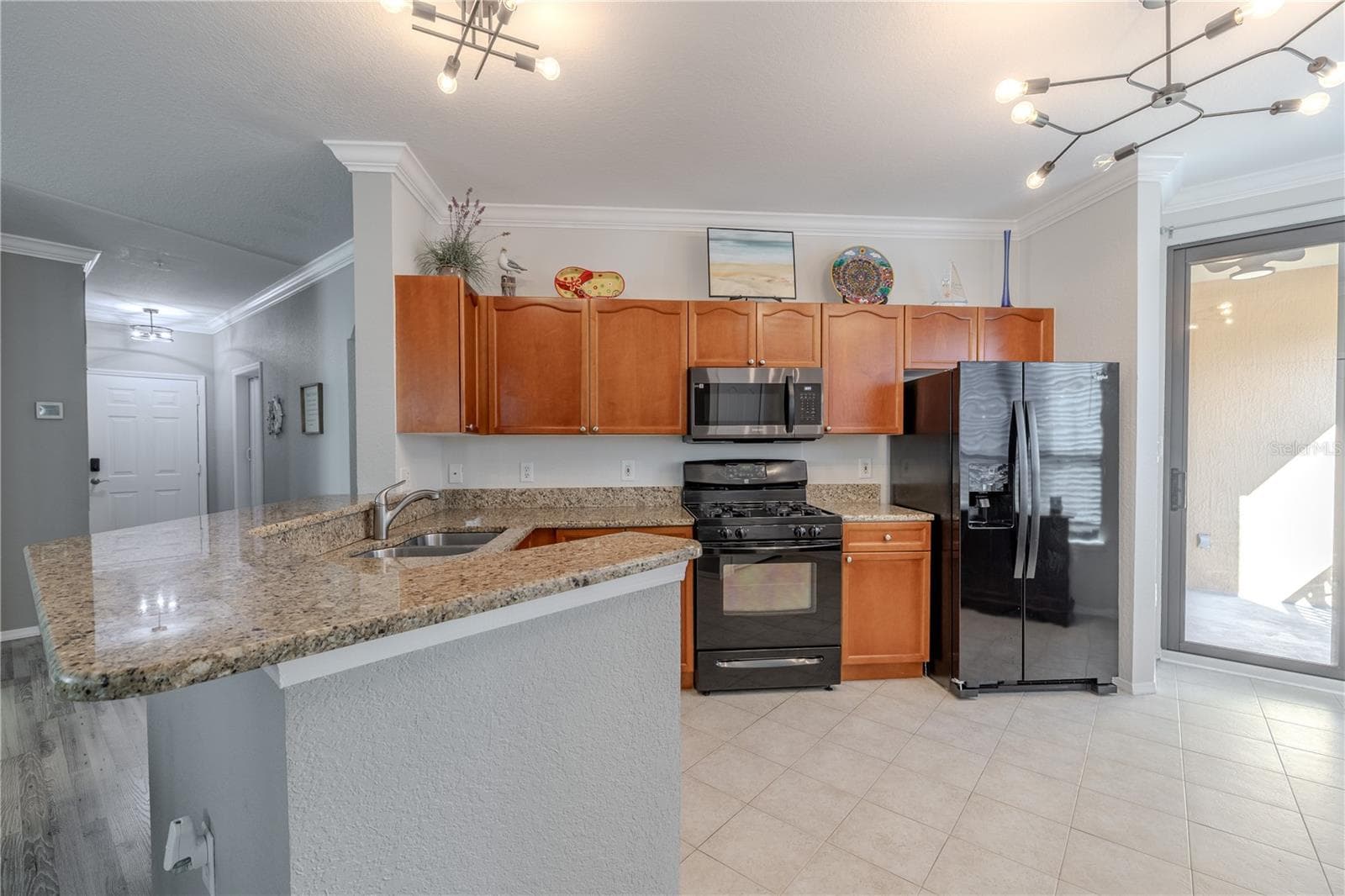 Open kitchen with a spacious granite breakfast bar, warm wood cabinetry, and black appliances, complemented by stylish modern lighting and an easy flow to the living area and screened lanai beyond. - 2738 VIA TIVOLI #220B, CLEARWATER