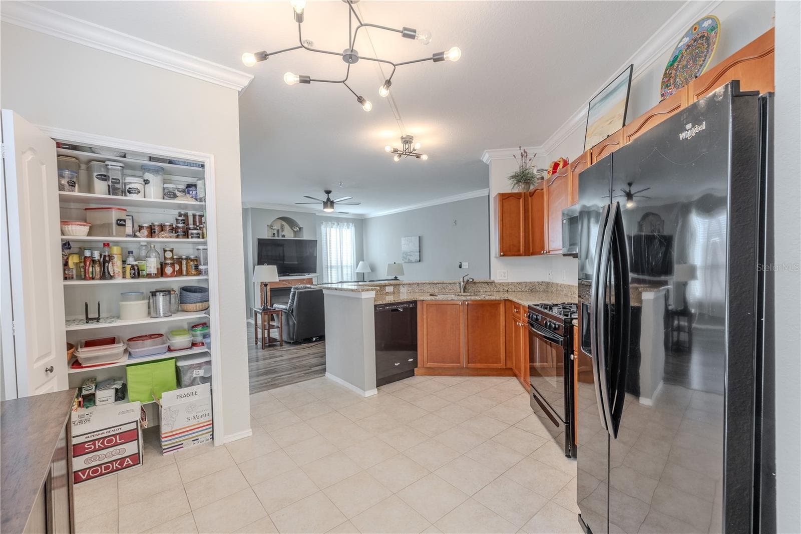 This kitchen view highlights the large pantry with double bi-fold doors, along with granite countertops, stainless appliances, warm wood cabinetry, and an open connection to the living area for seamless everyday living and entertaining. - 2738 VIA TIVOLI #220B, CLEARWATER