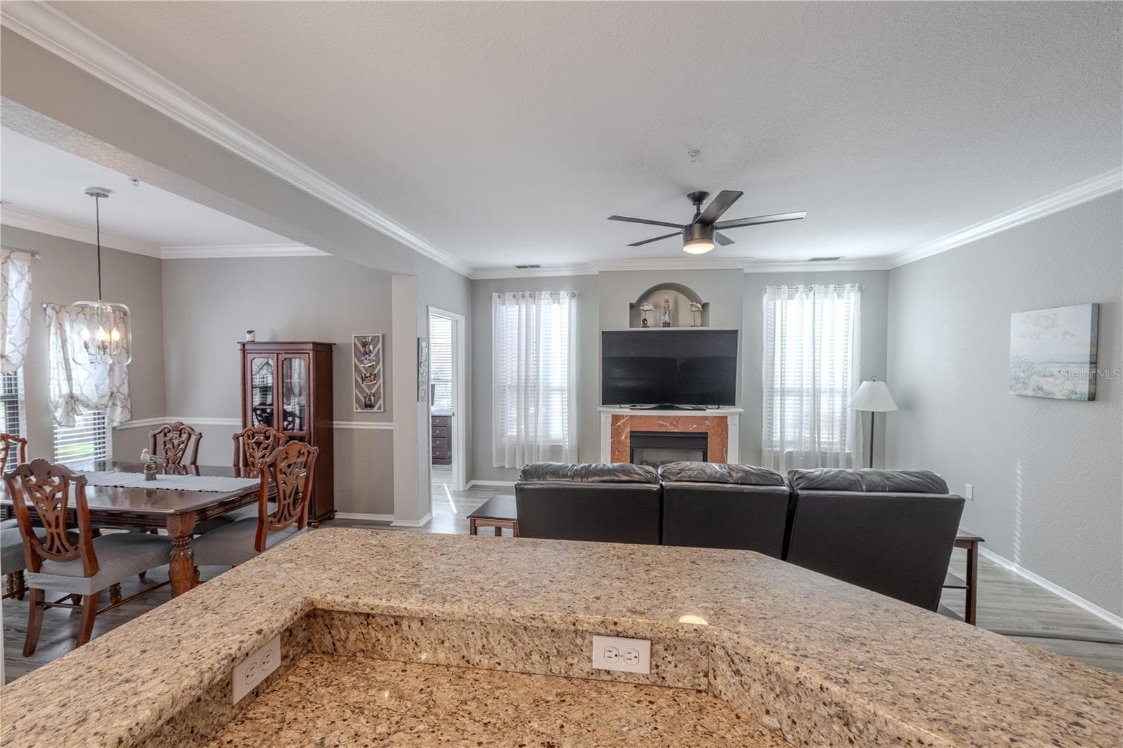 From the kitchen, the expansive granite counter overlooks the living and dining areas, highlighting the open layout, abundant natural light, and a cozy living room centered around a fireplace and modern ceiling fan. - 2738 VIA TIVOLI #220B, CLEARWATER