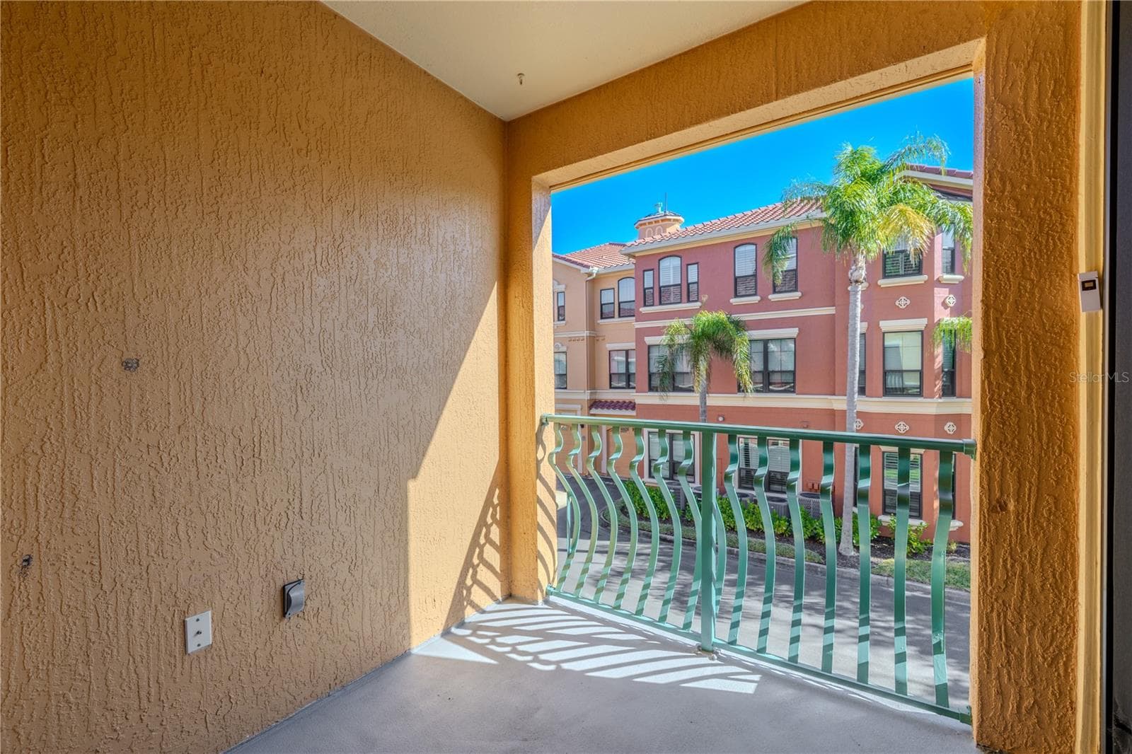 Private covered balcony primary bedroom view with sliding glass doors, offering a sunny outdoor space and elevated views of the palm-lined community streetscape. - 2738 VIA TIVOLI #220B, CLEARWATER