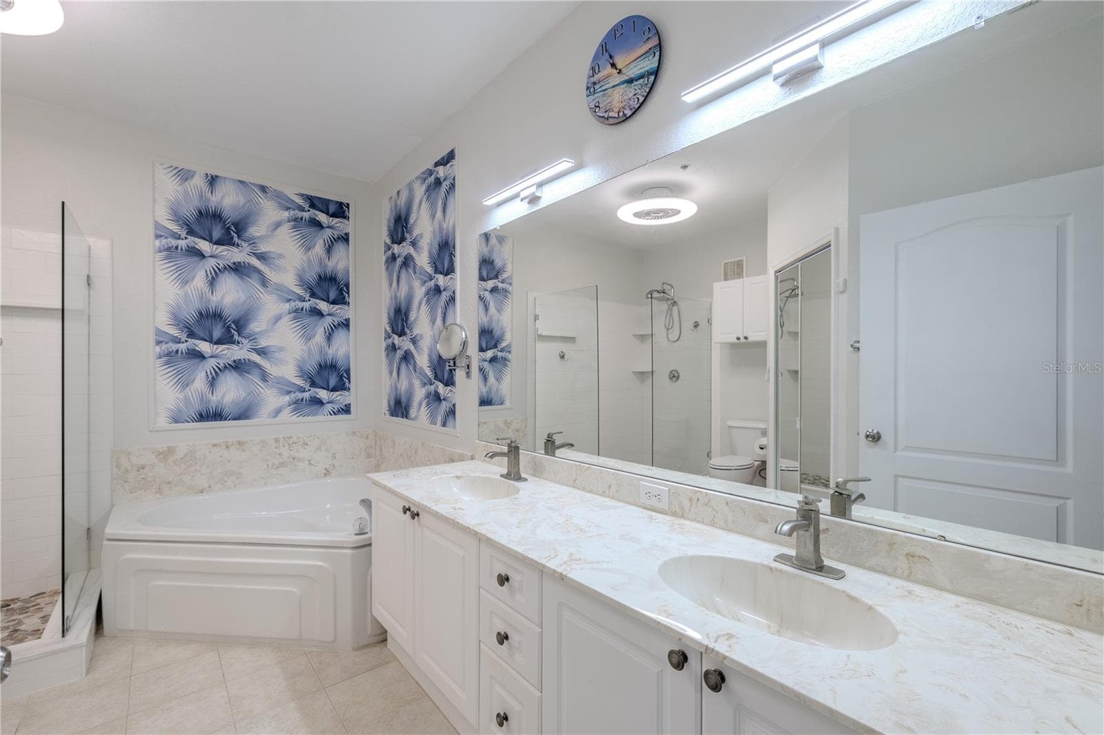 This view highlights the spa-inspired primary bathroom with a glimpse of the glass-enclosed walk-in shower featuring pebble-style flooring, a deep soaking tub with elegant tile surround, and bright, contemporary finishes throughout. - 2738 VIA TIVOLI #220B, CLEARWATER