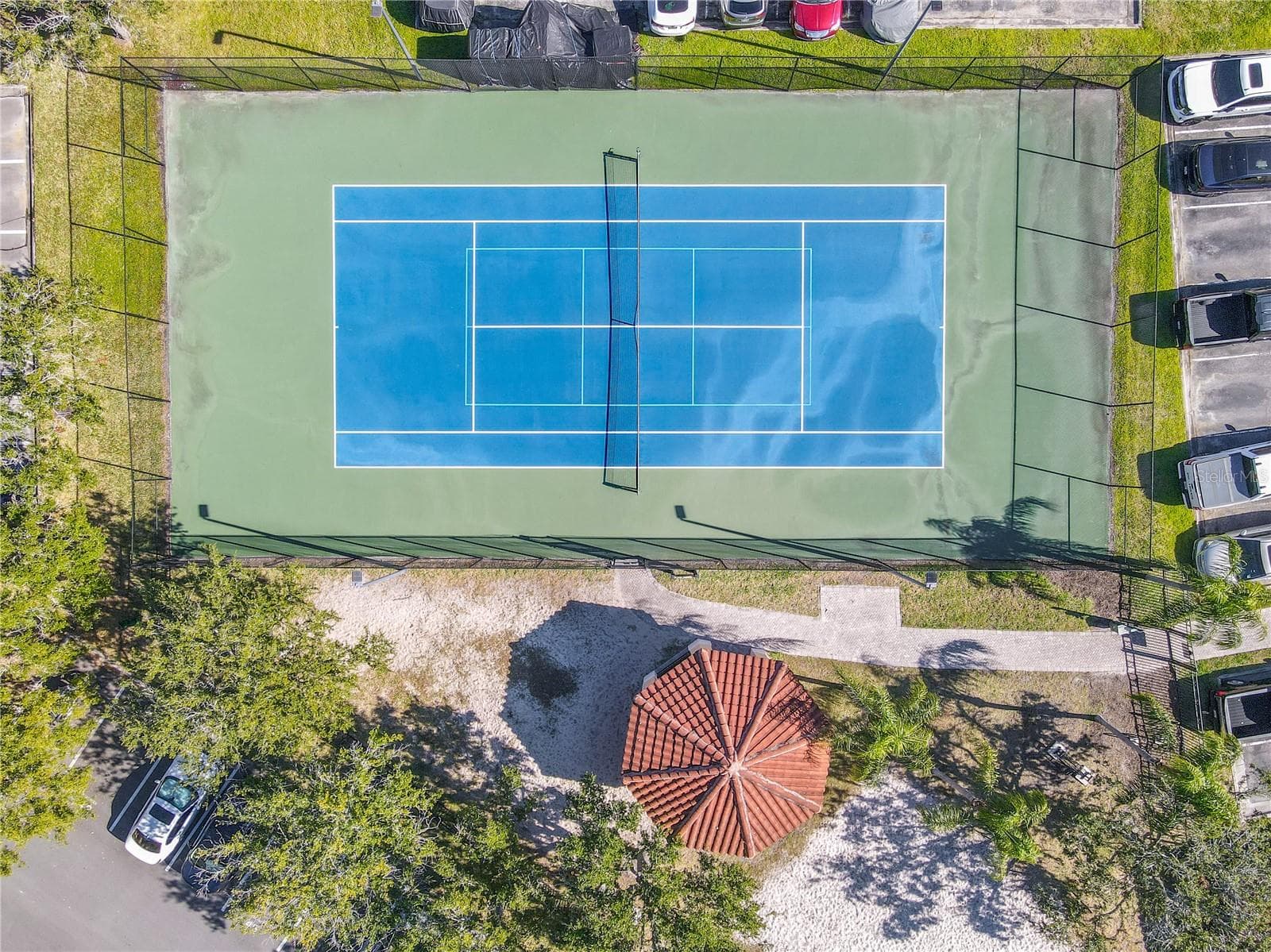 An aerial view highlighting the tranquil community with tennis courtyard with a shaded pavilion, sandy play area, and palm-lined green space, framed by elegant Mediterranean-style residences under clear blue skies. - 2738 VIA TIVOLI #220B, CLEARWATER