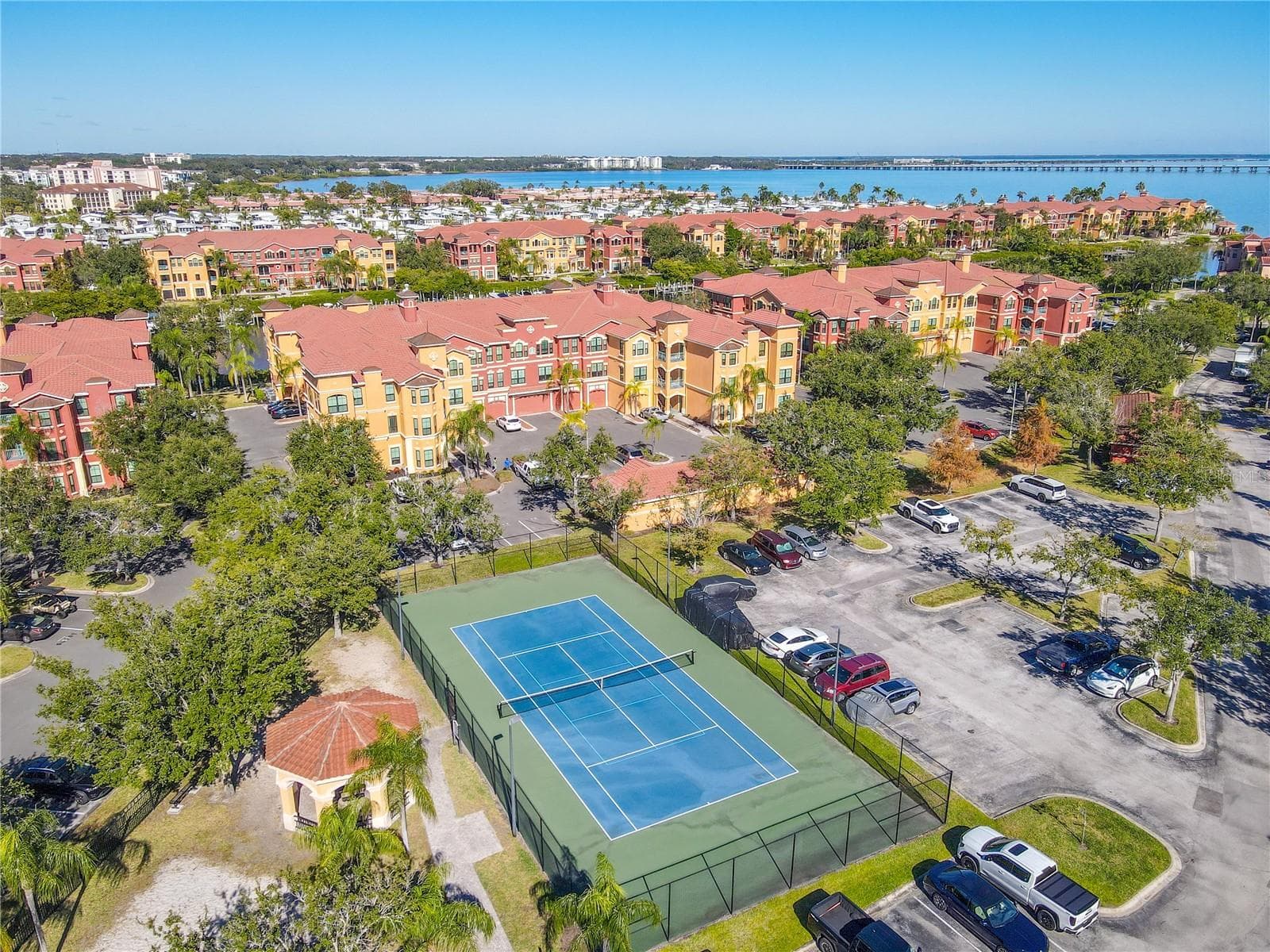 Another aerial view highlighting the tranquil community with tennis courtyard with a shaded pavilion, sandy play area, and palm-lined green space, framed by elegant water view of an the Mediterranean-style residences under clear blue skies. - 2738 VIA TIVOLI #220B, CLEARWATER