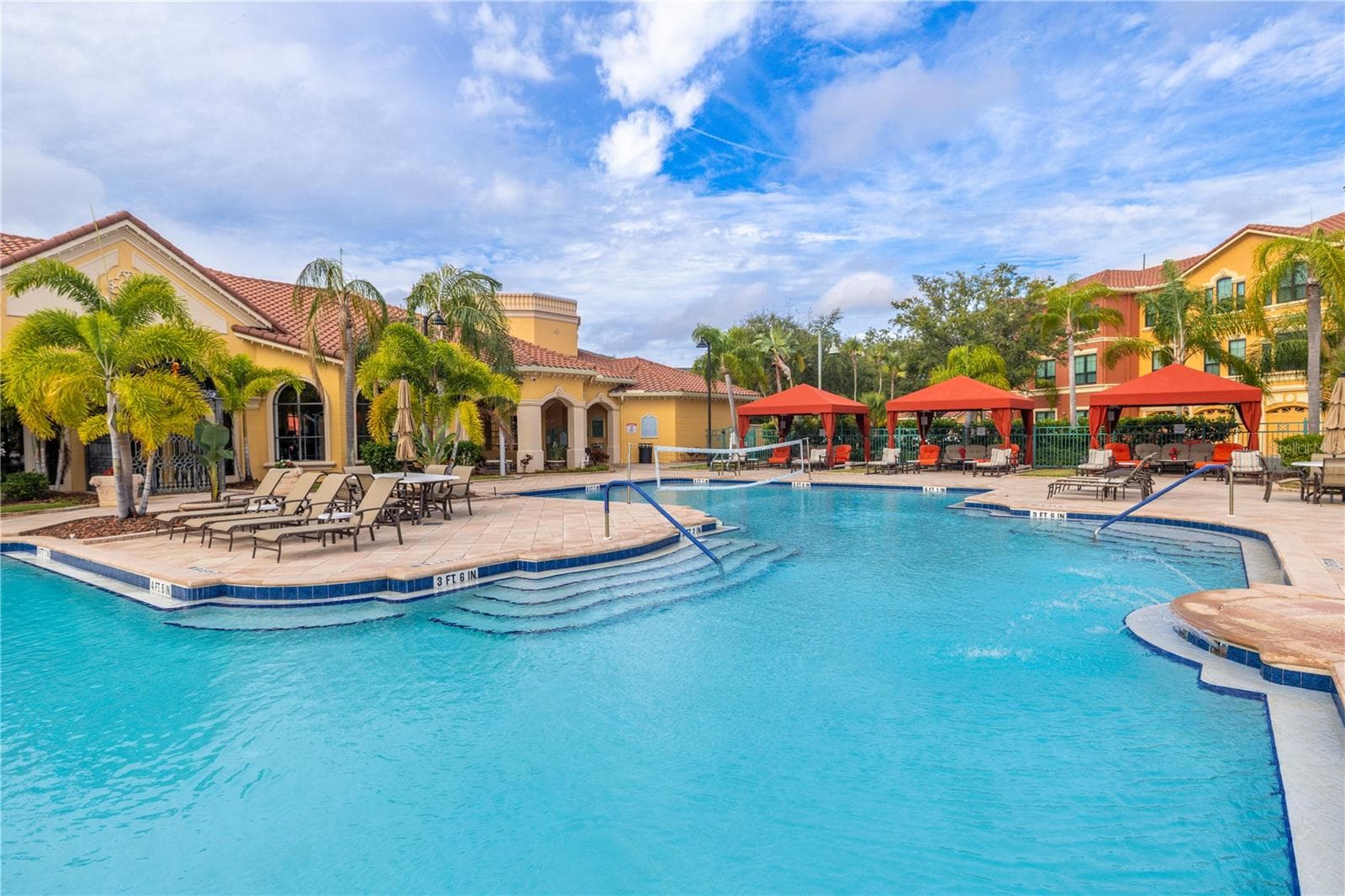 Resort-style clubhouse heated pool with a zero-entry sun shelf, palm-lined deck, lounge seating, and private cabanas—set against beautiful Mediterranean architecture for a true vacation-at-home vibe. - 2738 VIA TIVOLI #220B, CLEARWATER