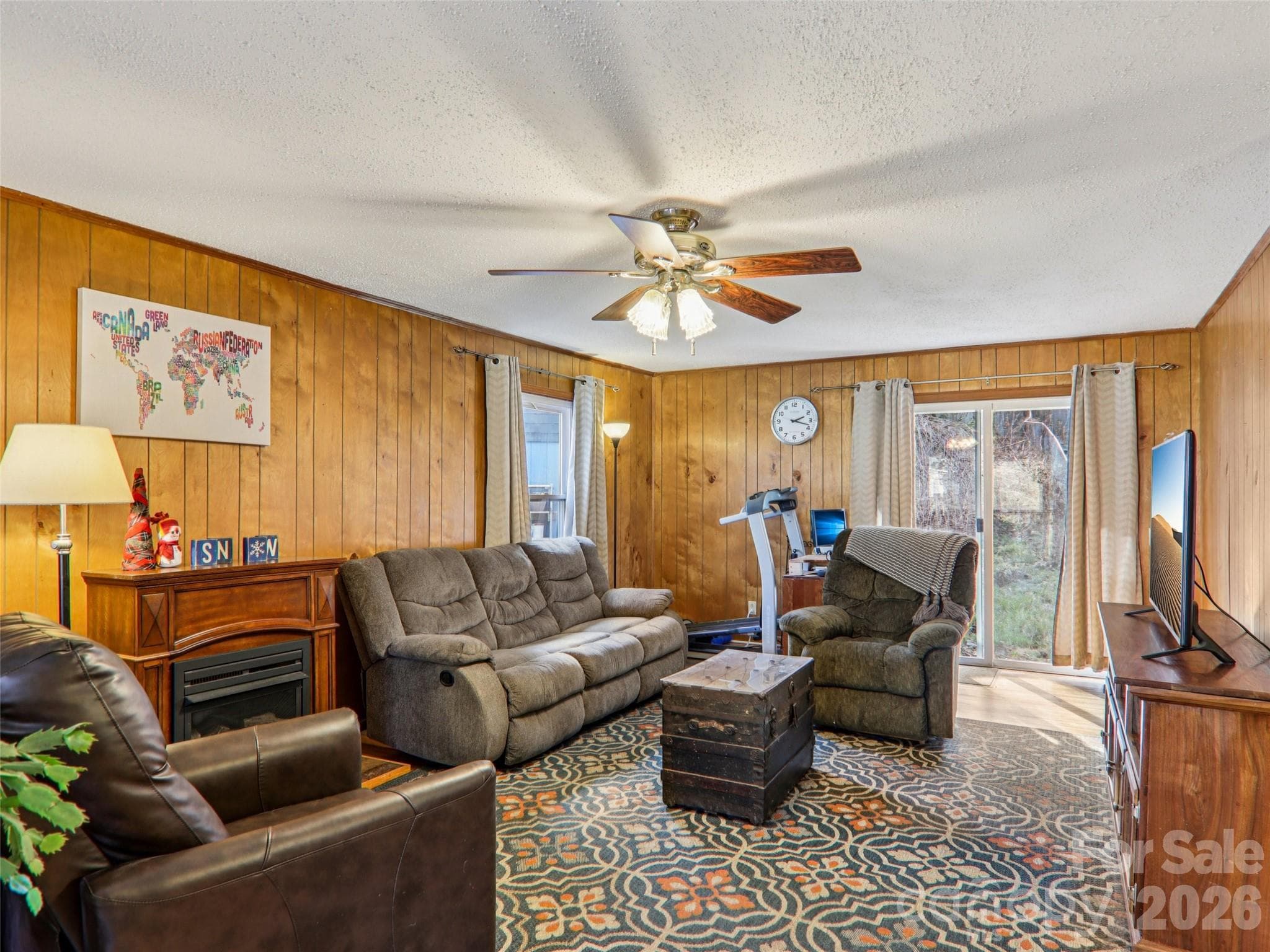 Comfortable living room with wood-paneled walls, ceiling fan, and generous natural light, featuring a non-vented propane fireplace that adds supplemental warmth and character to the space. - 7 Landon Mountain Lane, Weaverville