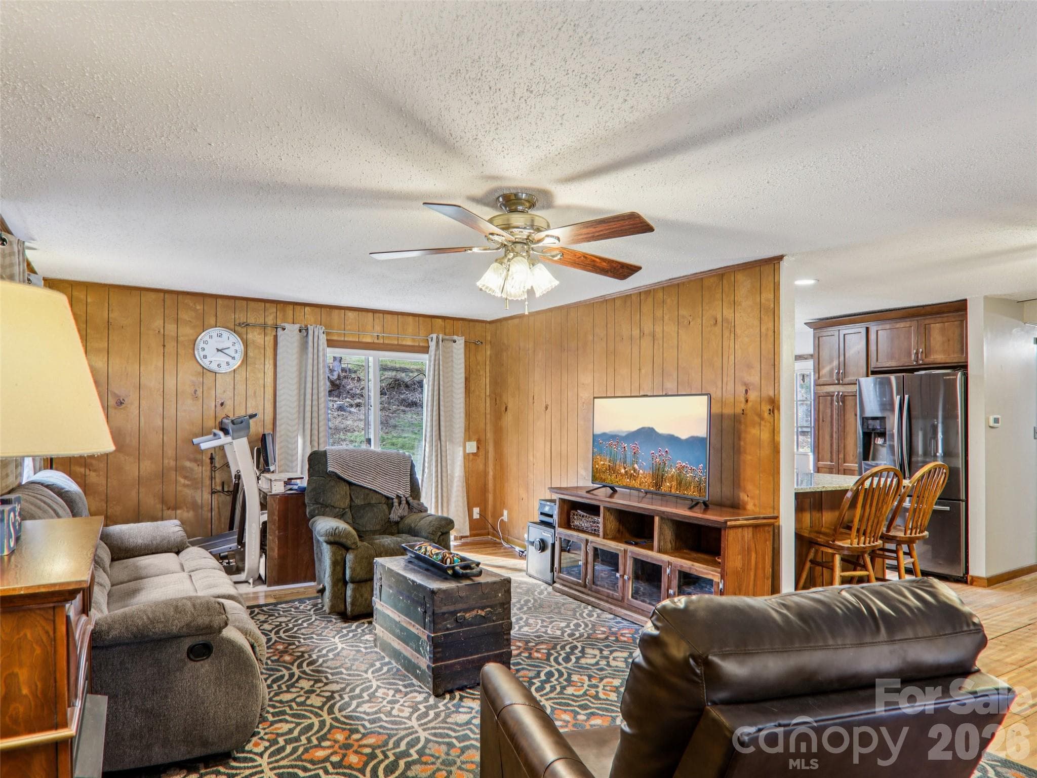 Alternate angle of the living room showing the open flow toward the kitchen, wood-paneled walls, ceiling fan, and natural light, creating a comfortable and functional main living space. - 7 Landon Mountain Lane, Weaverville