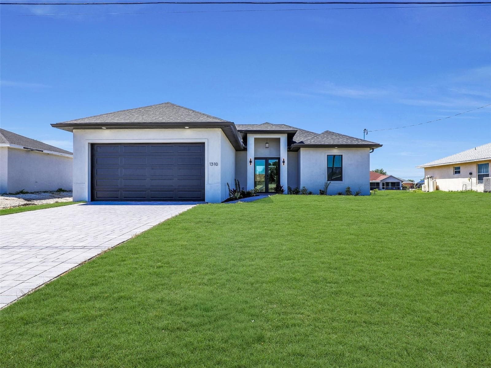Contemporary stucco exterior with a dark garage door and a contrasting paver drivewaythe color of the grass has been virtually enhanced - 1310 NW 11TH TER, CAPE CORAL