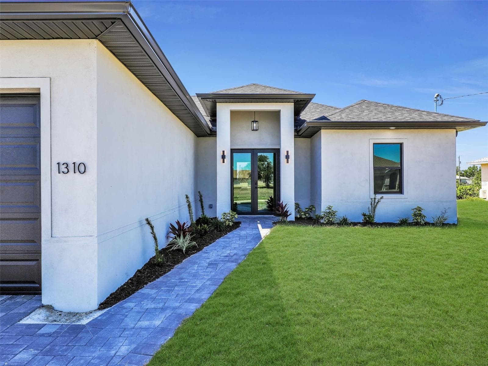 Contemporary stucco exterior with a covered entry, dark-framed double glass entry doors, a paver walkway, a manicured lawn, and professional landscaping the color of the grass has been virtually enhanced - 1310 NW 11TH TER, CAPE CORAL