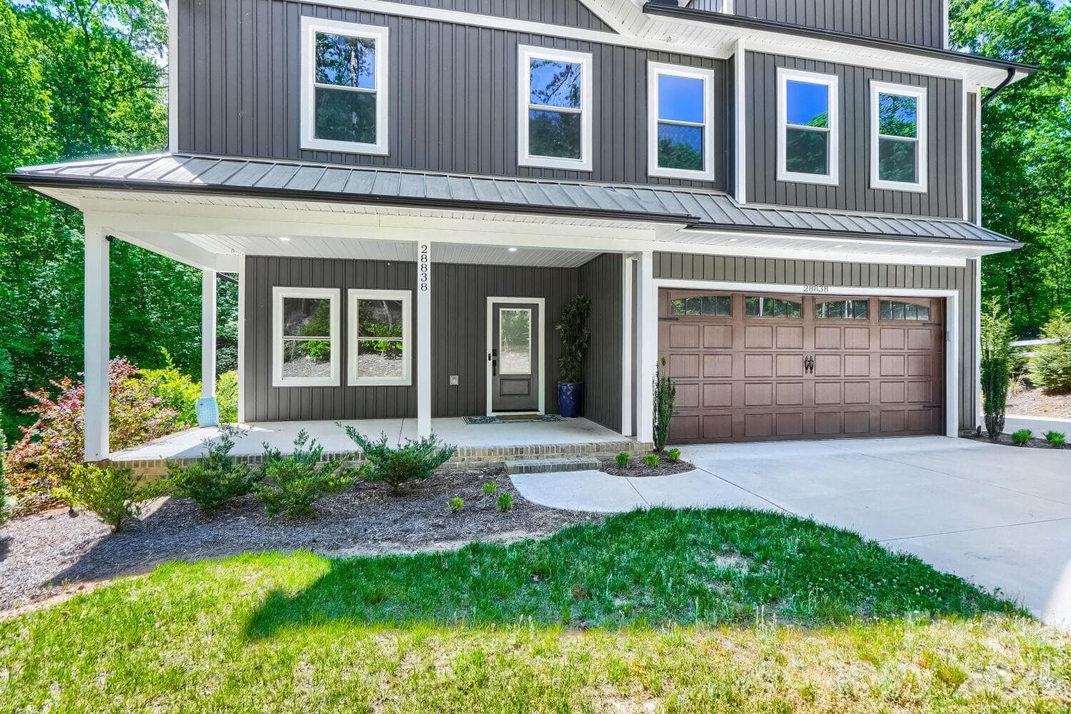 Charming covered front porch with board-and-batten exterior, standing seam metal roof accent, white trim details, and an attached two-car garage with carriage-style door — exceptional curb appeal set against a lush wooded backdrop. - Albemarle