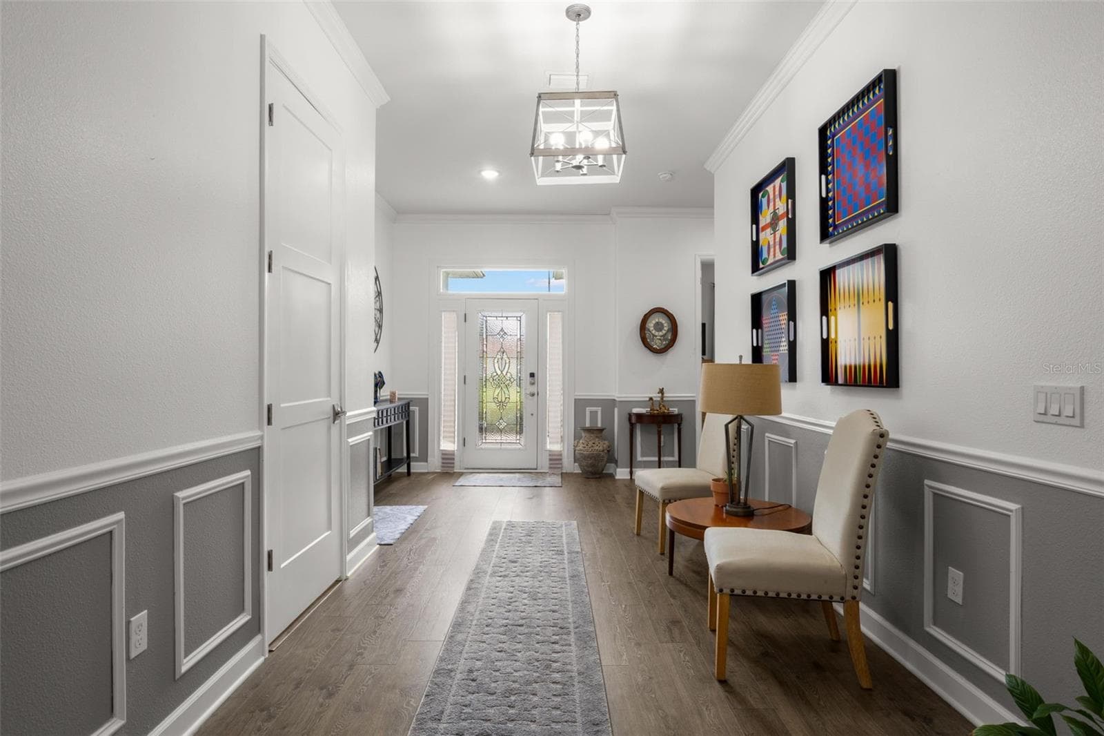 The impressive foyer showcases extensive crown molding, oversized base trim, wainscoting, chair rail detailing, recessed lighting, a statement chandelier, and wide-plank luxury vinyl flooring. A leaded glass entry door with sidelights and transom window adds natural light and architectural character to the welcoming entryway. - 1720 VERDE DR, CLEARWATER
