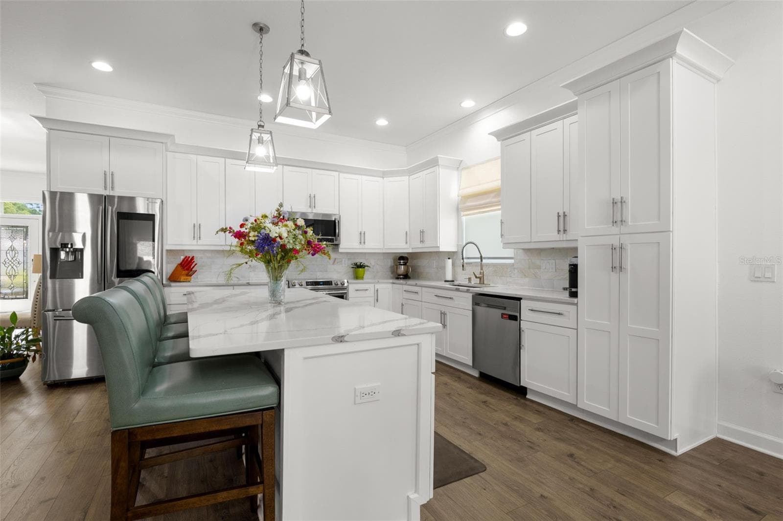 The designer kitchen features white shaker cabinetry with oversized crown molding, designer quartz countertops, a dramatic oversized center island with seating, upgraded stainless appliances, and an undermount farmhouse sink. Pendant lighting, recessed lighting, an onyx marble backsplash, and abundant cabinetry elevate both the functionality and visual appeal of the space. - 1720 VERDE DR, CLEARWATER