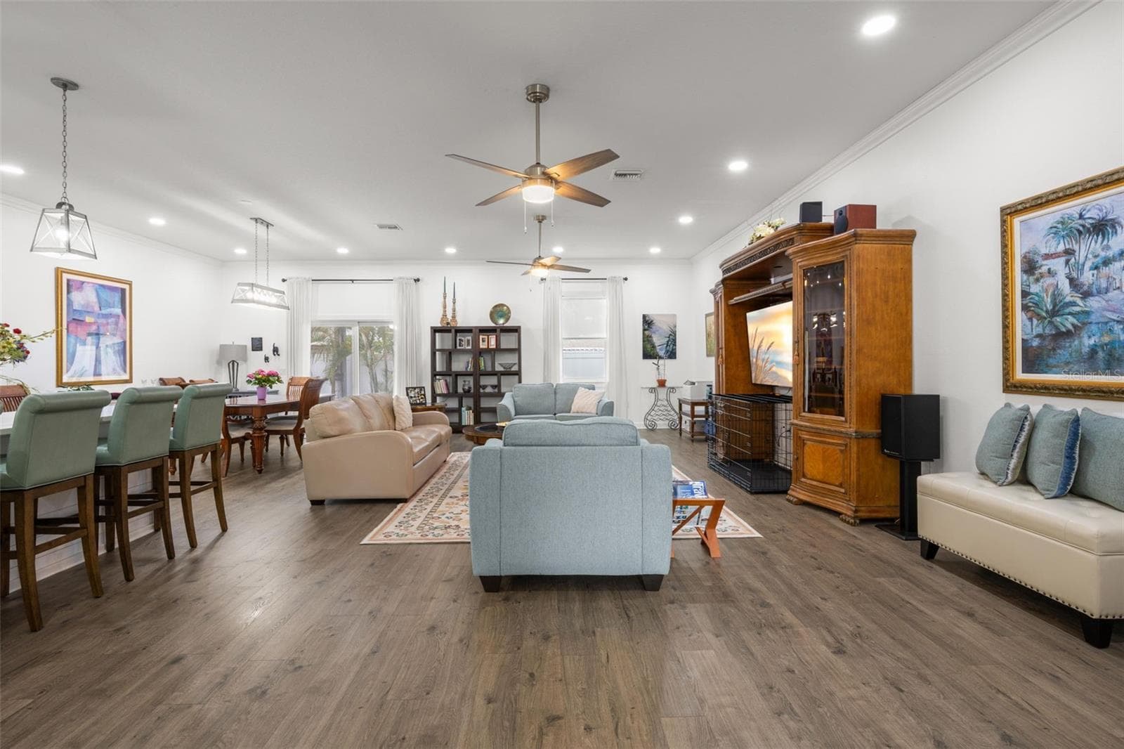 The spacious great room is anchored by wide-plank luxury vinyl flooring, crown molding, recessed lighting, and dual ceiling fans, creating a comfortable gathering space with clean sightlines to the kitchen and dining areas. Sliding glass doors and oversized windows contribute natural light throughout the open interior. - 1720 VERDE DR, CLEARWATER
