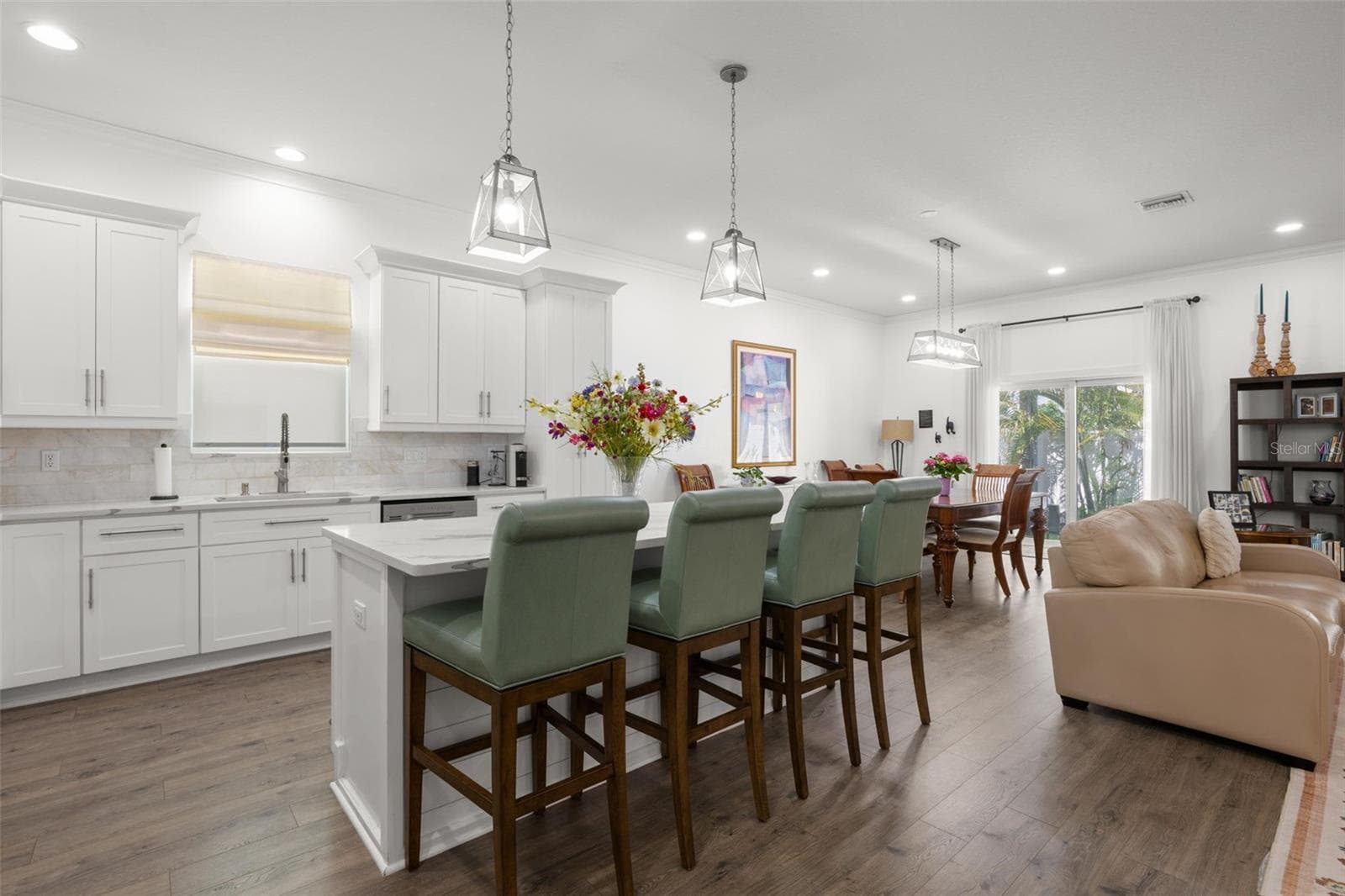 The oversized island offers generous seating and prep space beneath contemporary pendant lighting, while the surrounding open layout connects seamlessly to the dining and living areas. White shaker cabinetry with crown molding, designer quartz countertops, recessed lighting, and wide-plank luxury vinyl flooring continue throughout the bright, cohesive interior. - 1720 VERDE DR, CLEARWATER