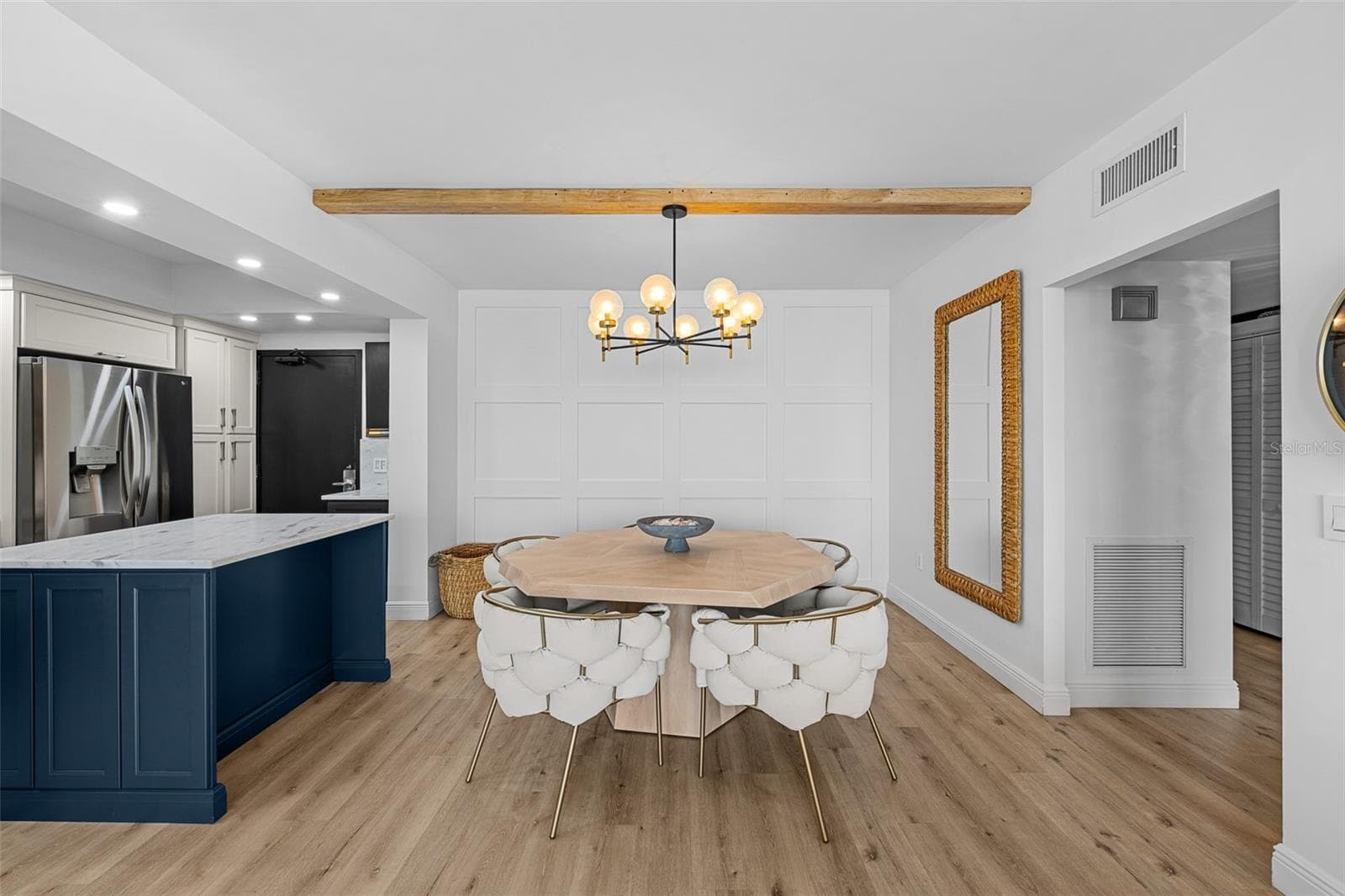 Dining area with a custom octagonal hardwood table, brass-and-glass chandelier, navy kitchen island in the background, and a panel-board accent wall. - 7100 SUNSHINE SKYWAY LN S #701, ST PETERSBURG