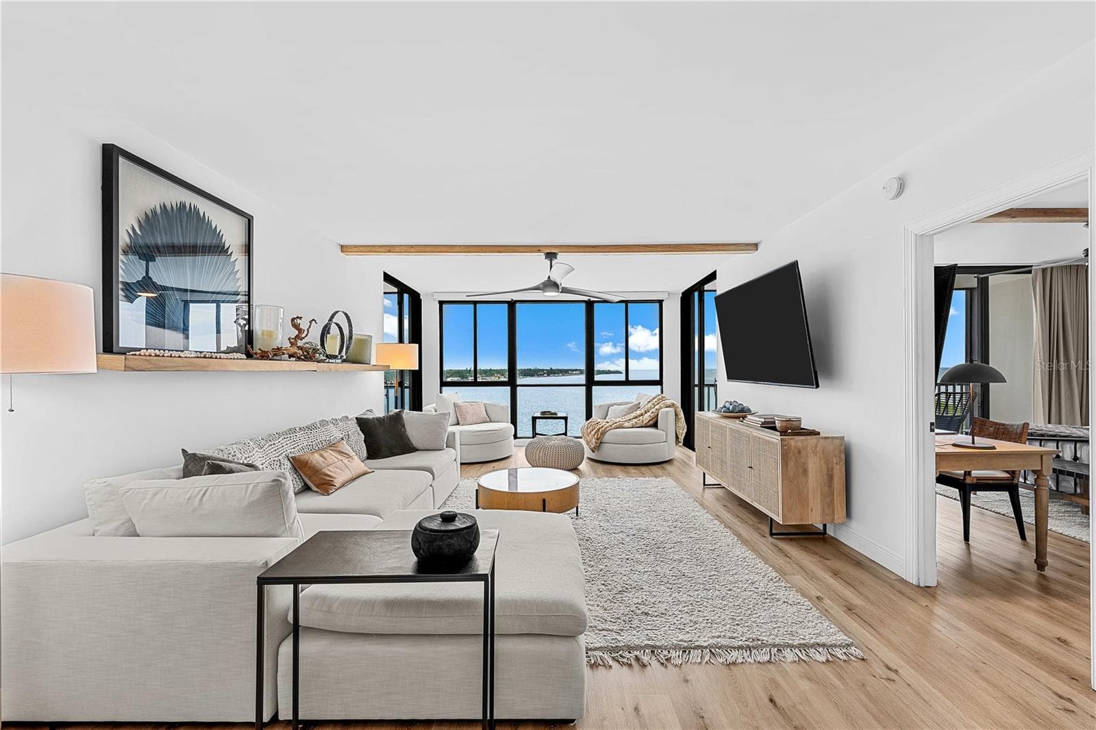 Open-concept living room with sectional seating, swivel lounge chairs, custom wood shelf, and panoramic Tampa Bay views through walls of glass. - 7100 SUNSHINE SKYWAY LN S #701, ST PETERSBURG