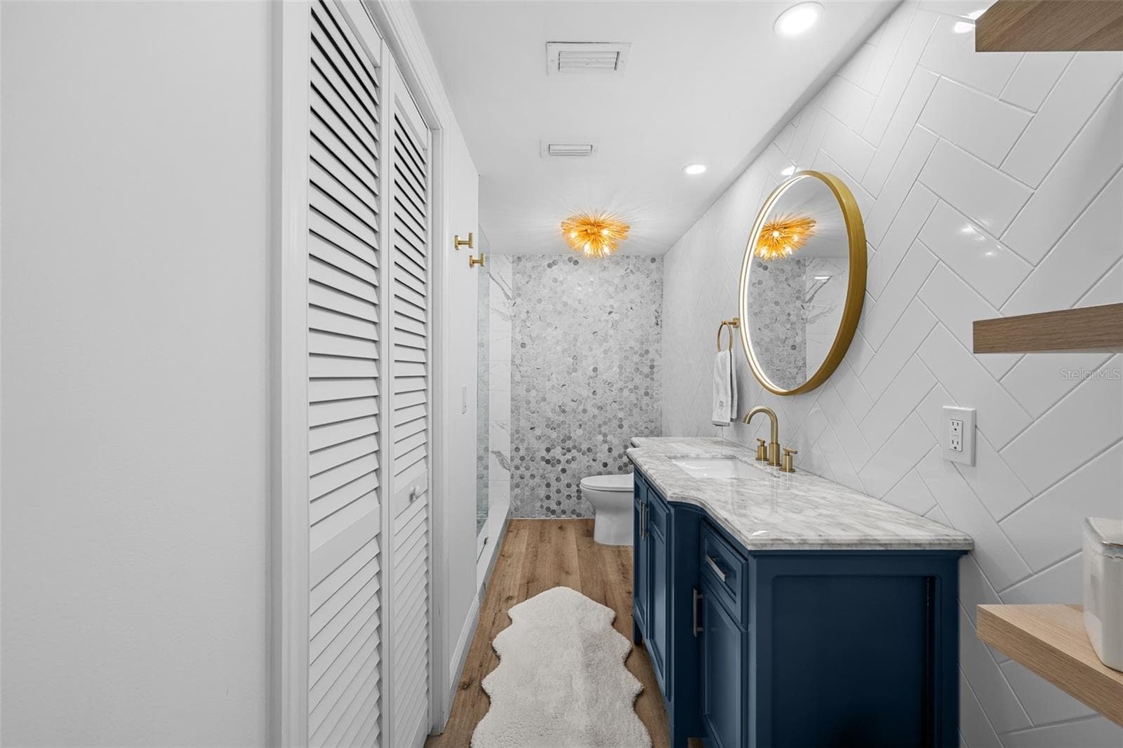 Renovated bath with herringbone white subway tile, a custom navy vanity, white marble counter, brass fixtures, and a walk-in marble shower. - 7100 SUNSHINE SKYWAY LN S #701, ST PETERSBURG