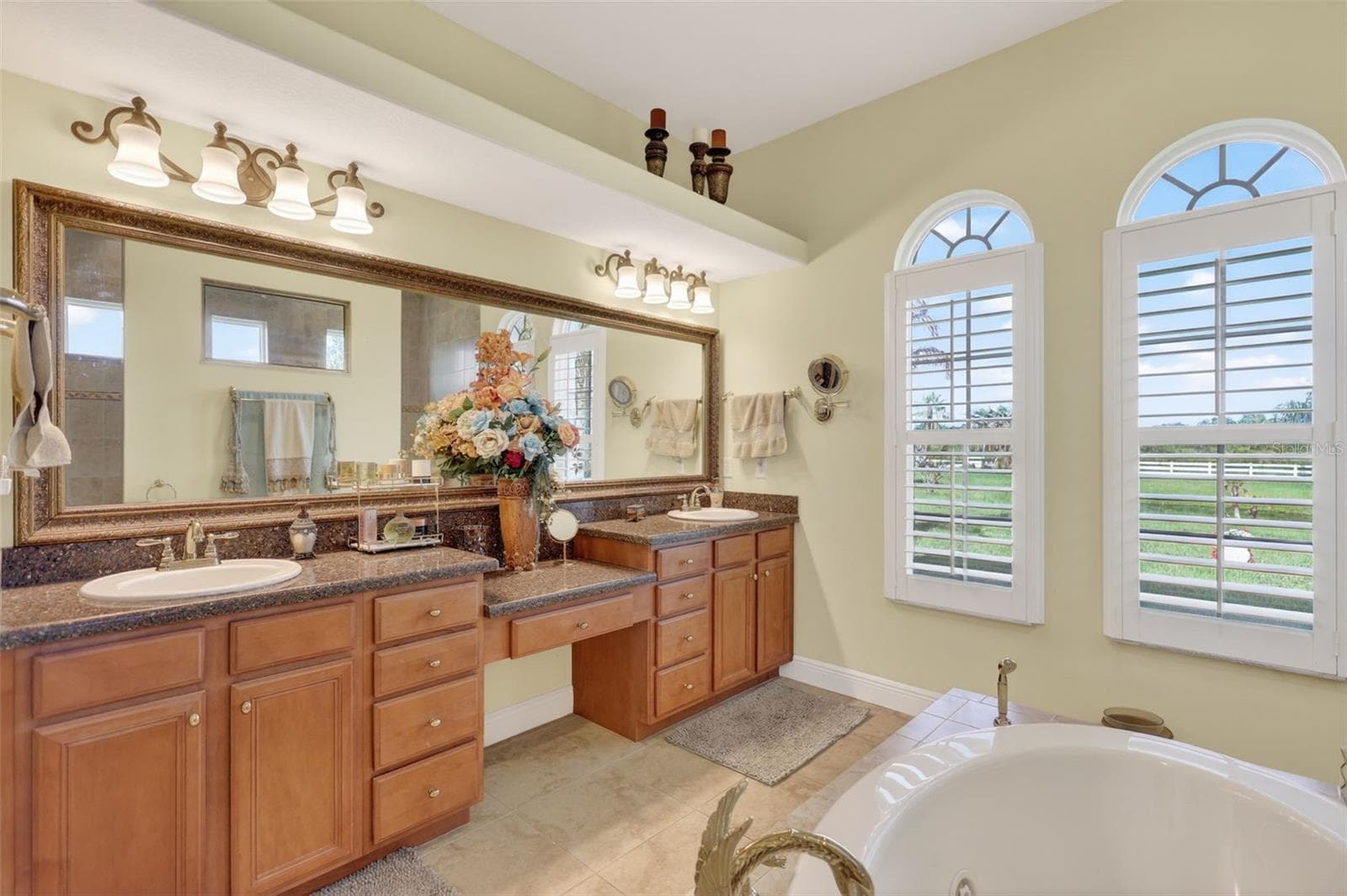 The elegant primary bath features dual vanities with abundant storage, a dedicated seated vanity area, and beautiful natural light from oversized windows. - 1210 408TH CT E, MYAKKA CITY