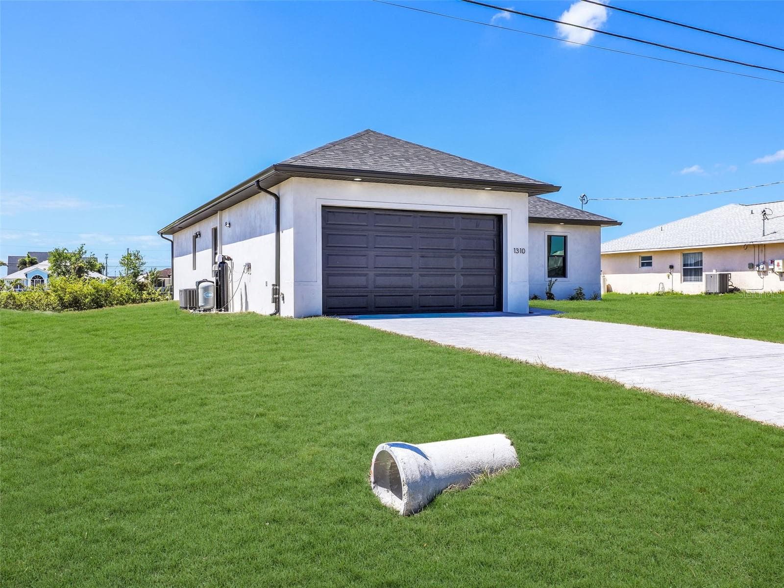 Contemporary single-story residence featuring a dark gray garage door, white exterior siding, and a dark shingle roof the color of the grass has been virtually enhanced - 1310 NW 11TH TER, CAPE CORAL