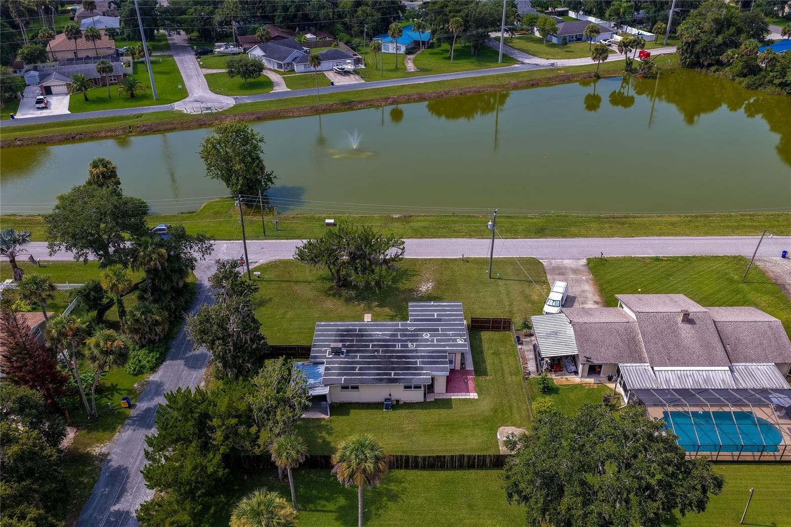 High-angle aerial view of the single-story home situated directly across from a peaceful neighborhood lake with a central water fountain. The spacious property features a grassy yard surrounded by mature palm trees and lush local vegetation. - 1101 16TH ST, EDGEWATER