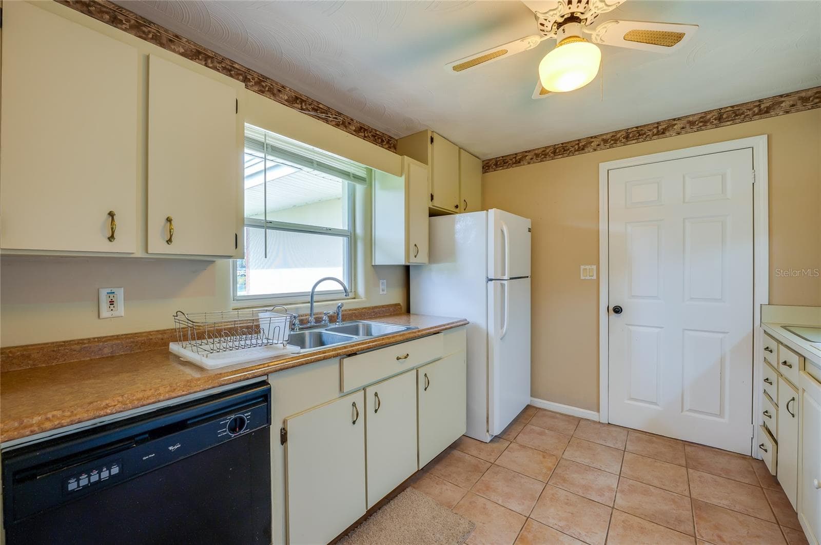 Kitchen work area showing a double basin sink, dishwasher, refrigerator, and neutral tiled flooring beneath an overhead ceiling fan. - 1101 16TH ST, EDGEWATER