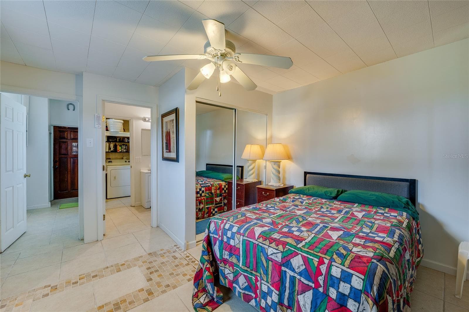 Tiled bedroom layout showcasing mirrored closet doors, a ceiling fan, and open doorway access toward the bathroom and utility spaces. - 1101 16TH ST, EDGEWATER