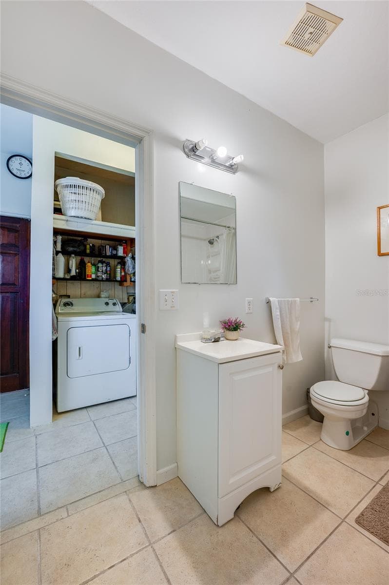 Bright bathroom layout showcasing a white pedestal-style vanity, toilet, tiled flooring, and a direct view into the adjoining laundry room. - 1101 16TH ST, EDGEWATER
