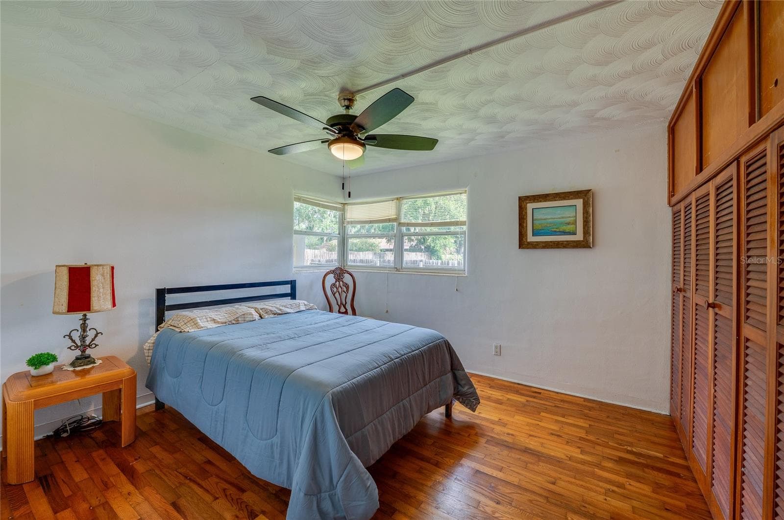 Bedroom area defined by warm hardwood flooring, a textured white ceiling with a fan, and full-height closet doors. Corner windows draw in natural light to brighten the layout. - 1101 16TH ST, EDGEWATER