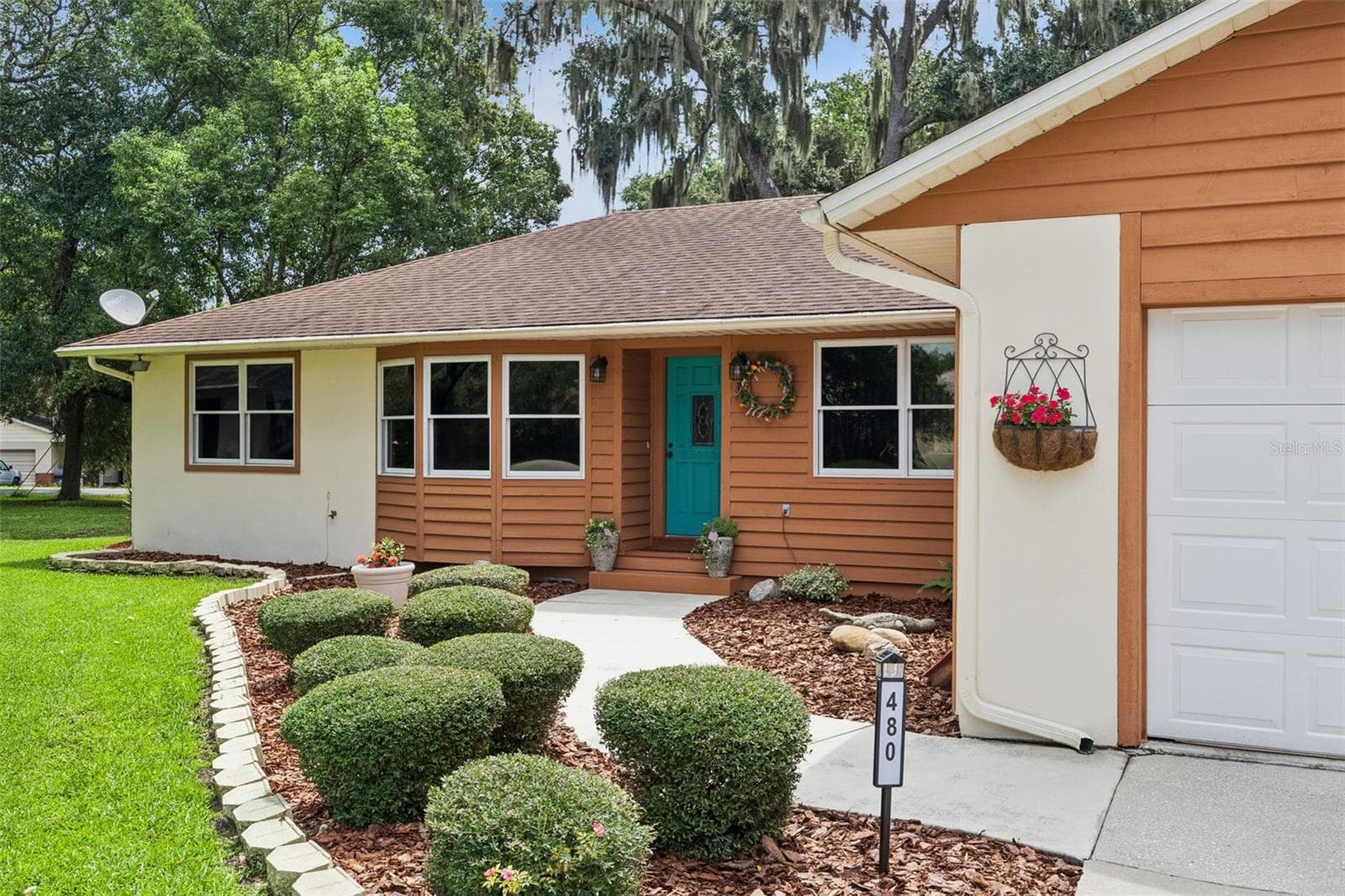 Inviting front entry at 480 Lake Forest Cir featuring a teal front door with wreath, manicured boxwood hedges, mulched flower beds, and hanging baskets alongside the 2-car garage in Mount Dora, FL - 480 LAKE FOREST CIR, MOUNT DORA