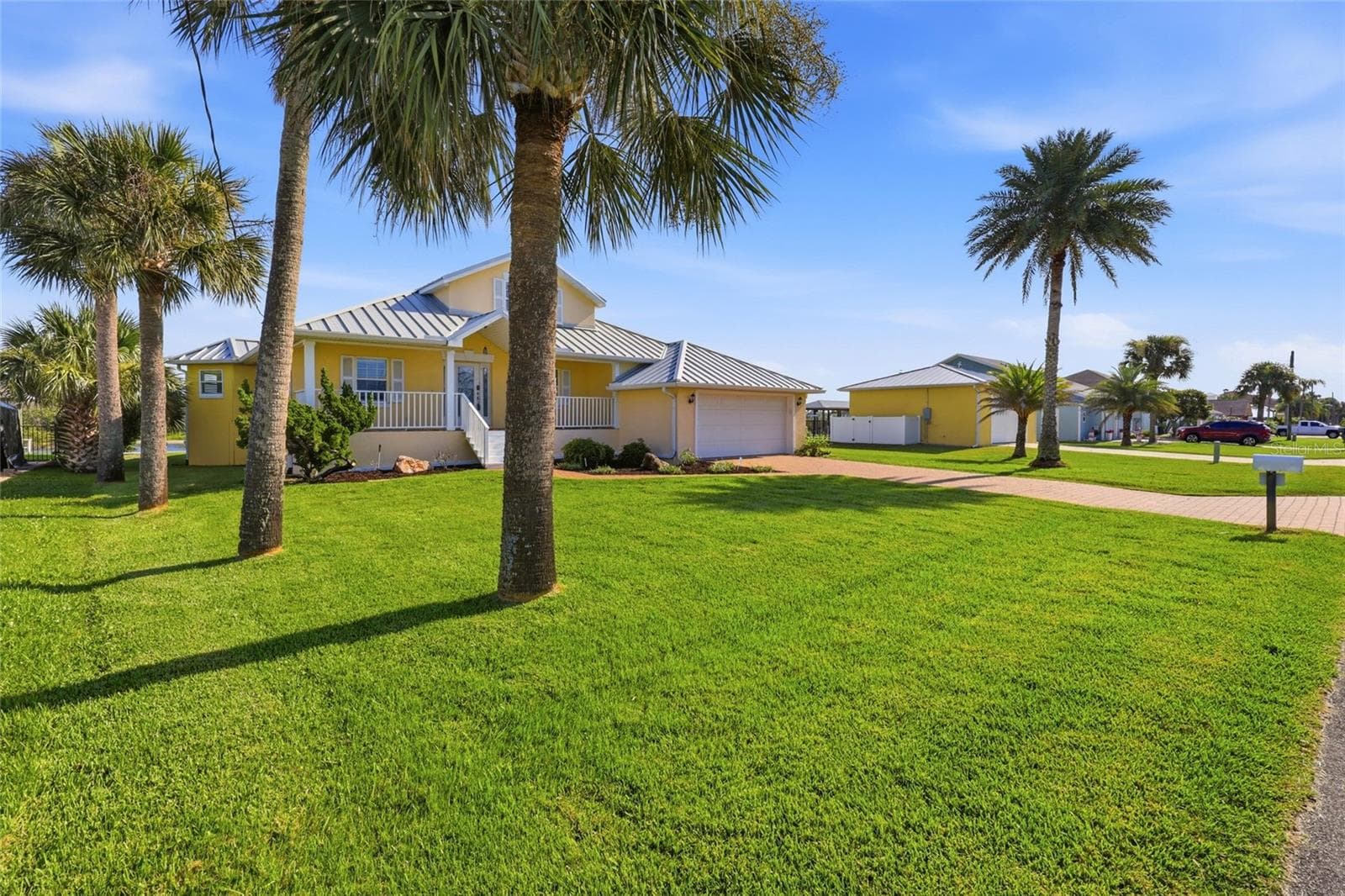 Front yard — Front yard view framed by mature palm trees, showing the yellow home's standing-seam metal roof, covered entry, and detached 2-car garage beyond the paver driveway. - 342 N 12TH ST, FLAGLER BEACH