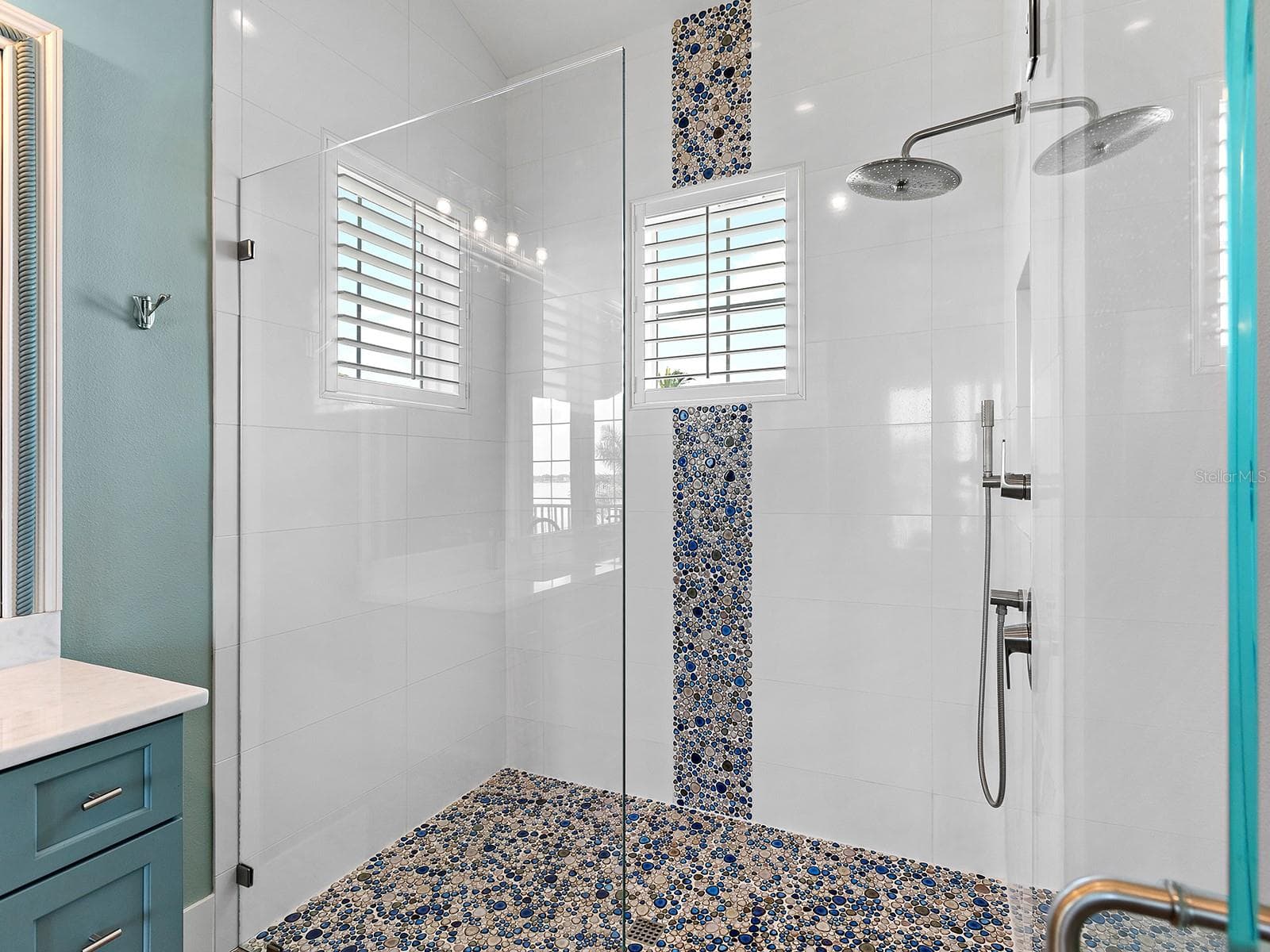 Luxe walk-in shower oasis with a vibrant blue and tan river rock mosaic feature wall, matching pebbled floor, a wall-mounted showerhead, and privacy-enhancing plantation shutters - 4024 11TH STREET CT W, PALMETTO