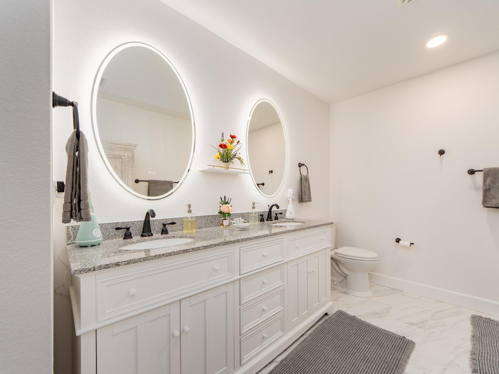 Spa-inspired primary en suite bathroom featuring a long double vanity, statement LED backlit mirrors, sleek fixtures, generous counter space, recessed lighting, and bright, large-format tile flooring for a clean, modern feel. - 1207 TIMBERBROOKE DR, PALM HARBOR