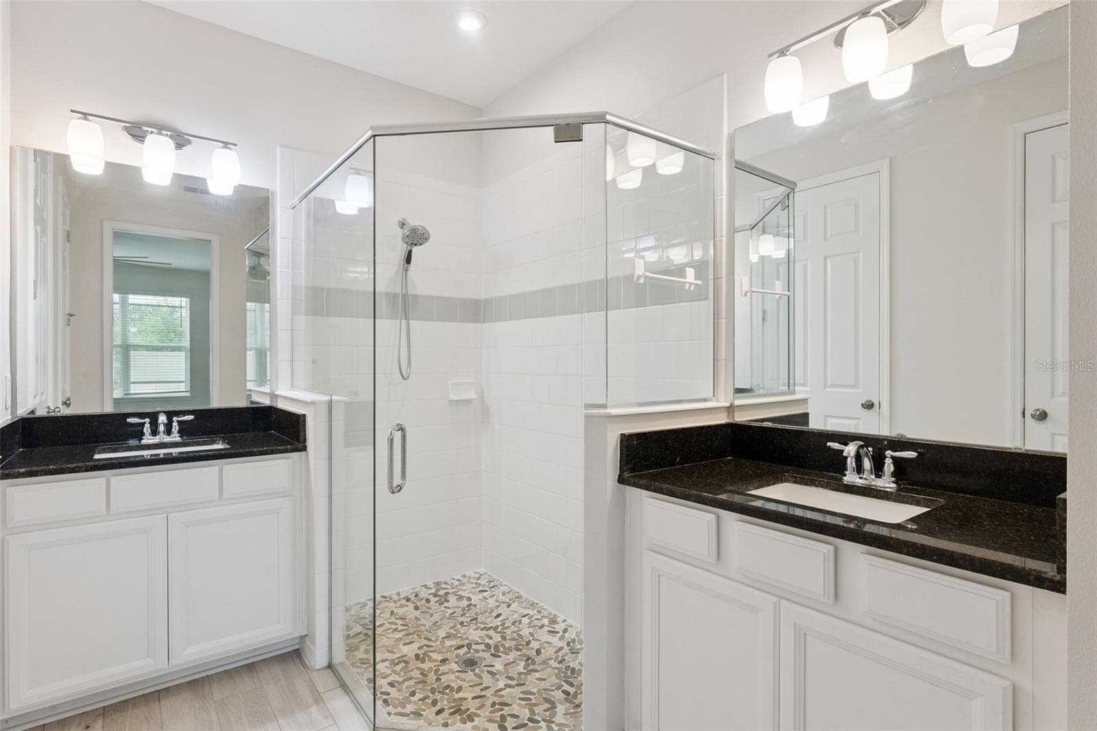 Downstairs Primary Bathroom – Bright ensuite bath features dual vanities with dark stone countertops, abundant white cabinetry, oversized mirrors, and a glass-enclosed tiled shower with a decorative pebble-style floor. - 1287 VERDANT GLADE PL, WINTER PARK
