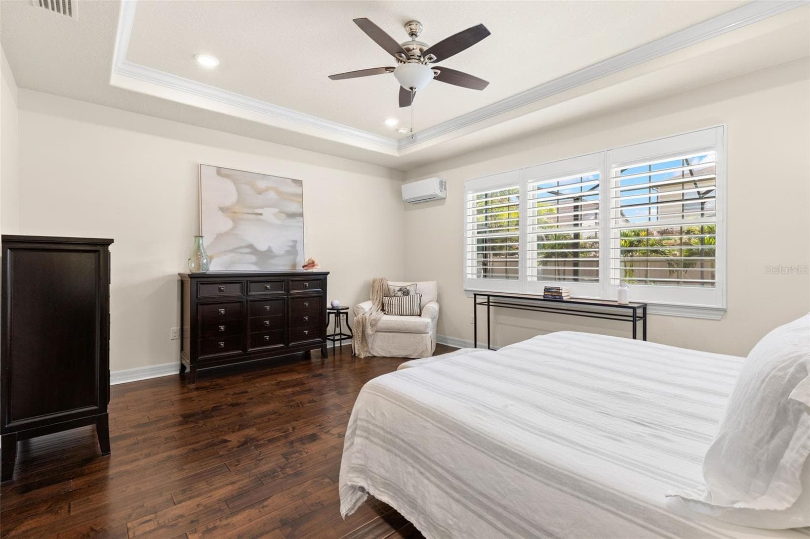 A warm, neutral palette gives the primary bedroom a calm, polished feel, with soft cream walls and crisp trim set against rich, dark engineered hardwood floors. Plantation shutters, a tray ceiling with crown molding, and a ceiling fan complete the comfortably elegant space. - 1528 FOREST GLEN CT, PALM HARBOR