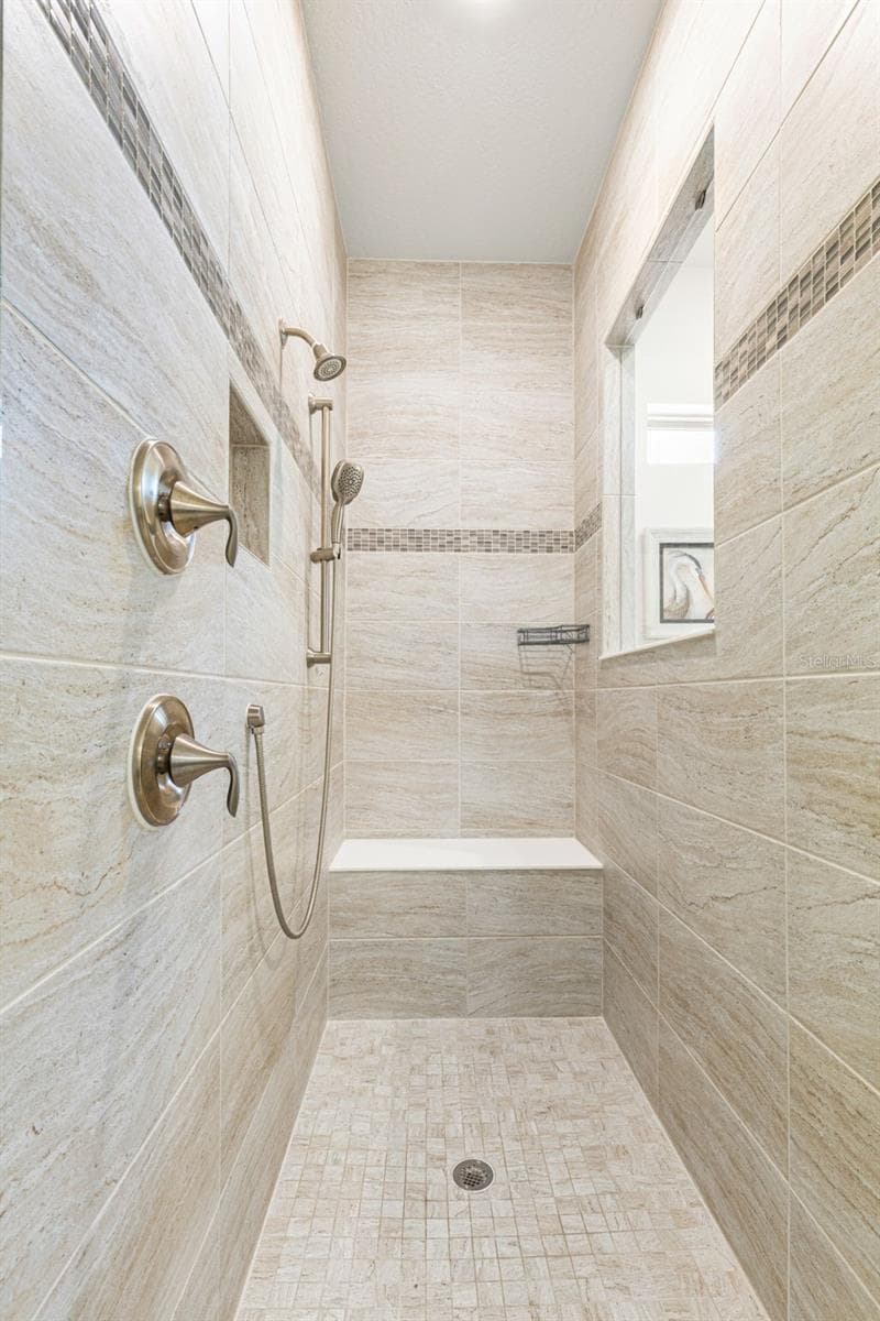 The oversized walk-in shower is finished from floor to ceiling in coordinating tile, with a built-in bench, recessed niche, decorative accent band, fixed showerhead and handheld spray, a spacious, thoughtfully designed complement to the primary bath. - 1528 FOREST GLEN CT, PALM HARBOR