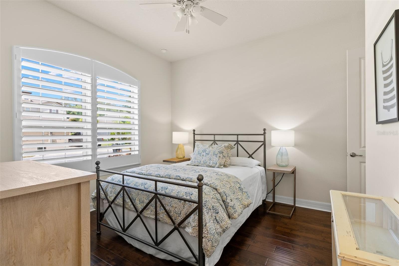 Bedroom 2 begins the home’s separate secondary-bedroom wing, with rich engineered hardwood flooring, a ceiling fan and plantation shutters framing the arched window. Neutral finishes keep the room light and adaptable, with plenty of space for a complete bedroom setup. - 1528 FOREST GLEN CT, PALM HARBOR